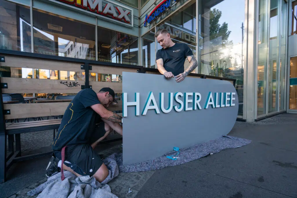 Zwei Arbeiter montieren ein Schild mit der Aufschrift 'HAUSER ALLEE' vor den Schönhauser Allee Arcaden. Einer kniet auf einer Decke und arbeitet an der Unterseite, während der andere das Schild hält. Im Hintergrund sind ein Cafébereich und Glasfassaden zu sehen.