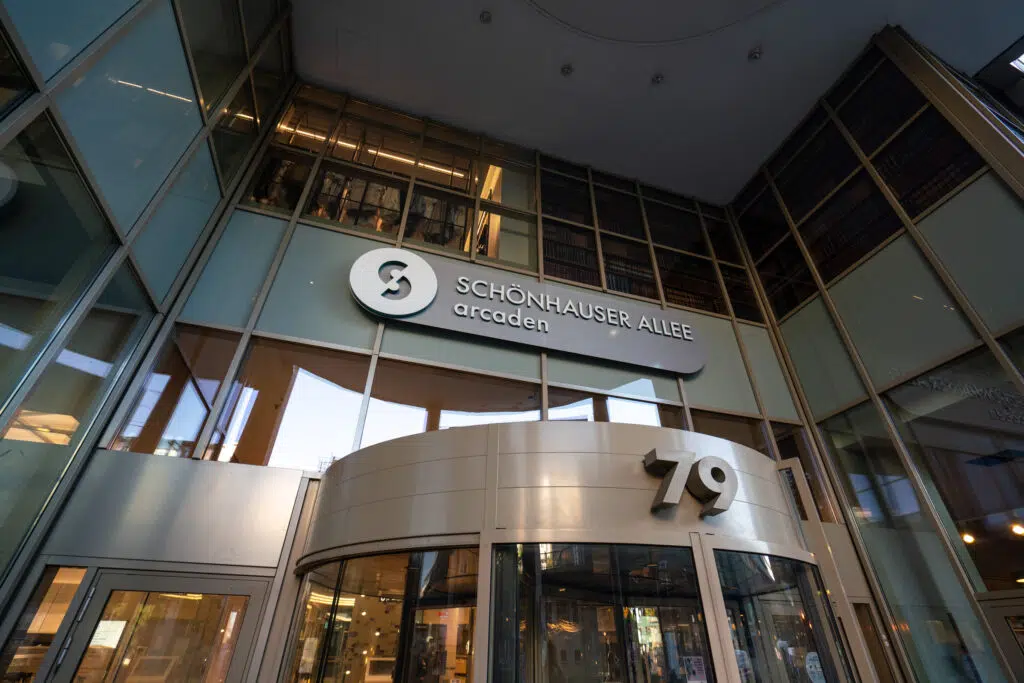 Eingangsbereich der Schönhauser Allee Arcaden mit moderner Glasfassade. Über der runden Drehtür mit der Hausnummer 79 befindet sich das Logo der Einkaufspassage. Reflexionen in den Fenstern zeigen die Umgebung. Innenbeleuchtung strahlt durch die Glasflächen.