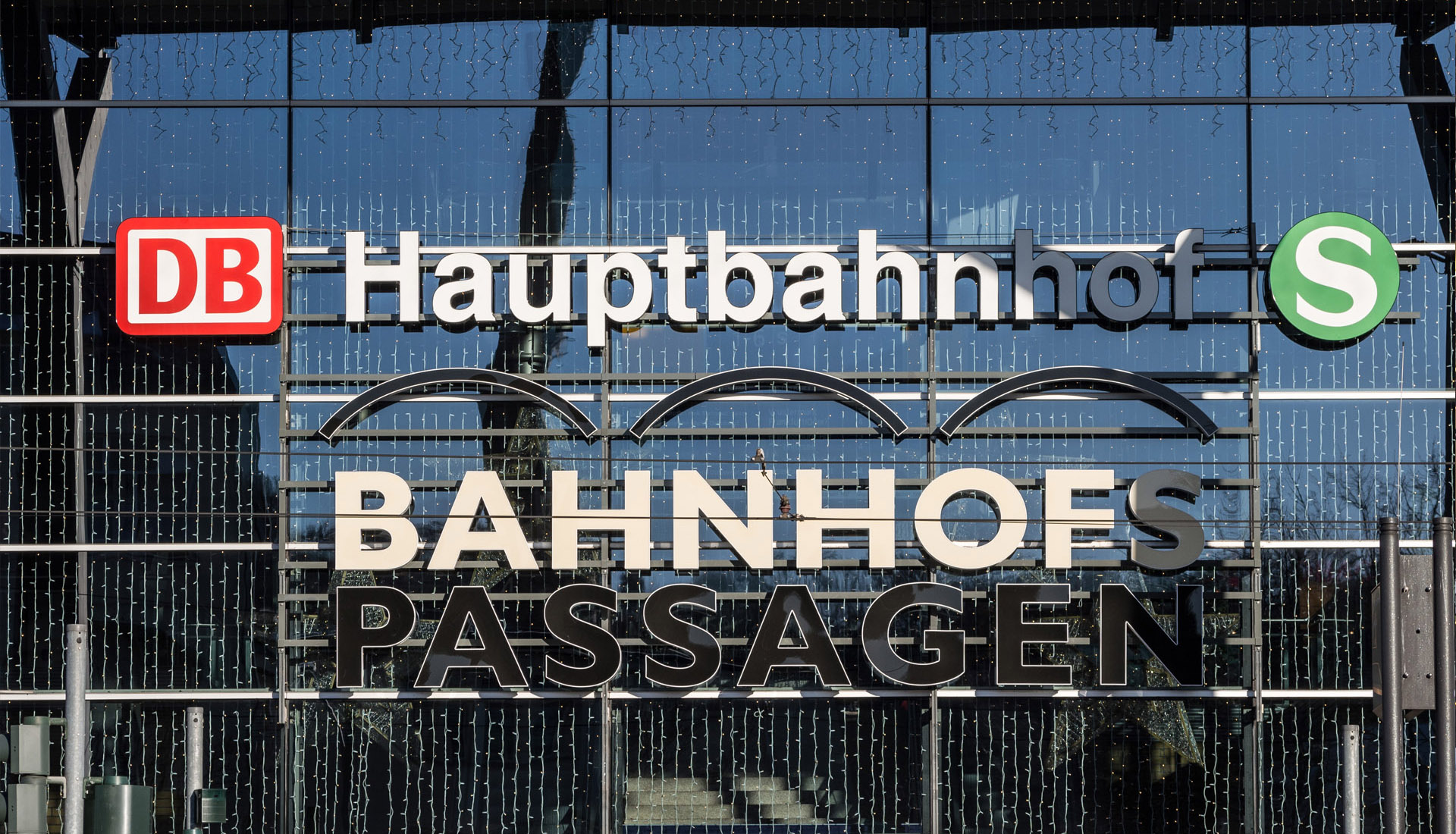 Frontansicht der Glasfassade des Bahnhofs mit Schriftzügen „DB Hauptbahnhof S“ und „Bahnhofspassagen“. Logos der Deutschen Bahn und S-Bahn sind deutlich sichtbar, im Hintergrund spiegeln sich Strukturen des Bahnhofs und der Himmel.