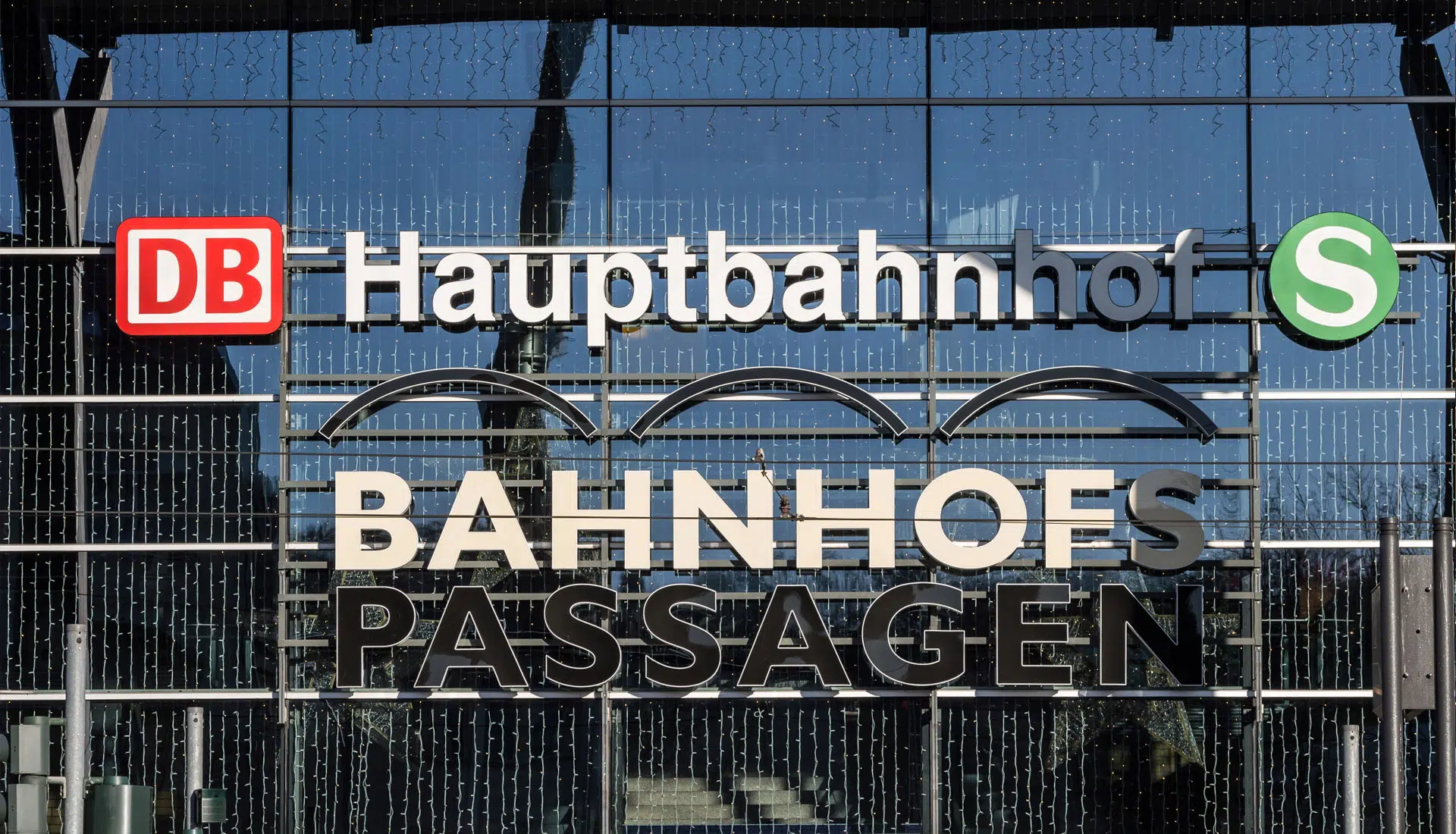 Frontansicht der Glasfassade des Bahnhofs mit Schriftzügen „DB Hauptbahnhof S“ und „Bahnhofspassagen“. Logos der Deutschen Bahn und S-Bahn sind deutlich sichtbar, im Hintergrund spiegeln sich Strukturen des Bahnhofs und der Himmel.
