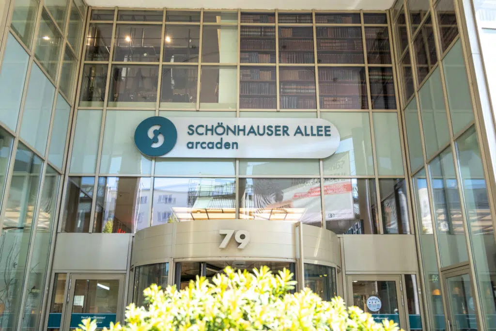 Der Eingangsbereich der Schönhauser Allee Arcaden mit einer Glasfassade und dem neuen, modern gestalteten Logo oberhalb der Drehtür. Die Hausnummer '79' ist über dem Eingang angebracht, reflektierende Fenster zeigen die Umgebung.