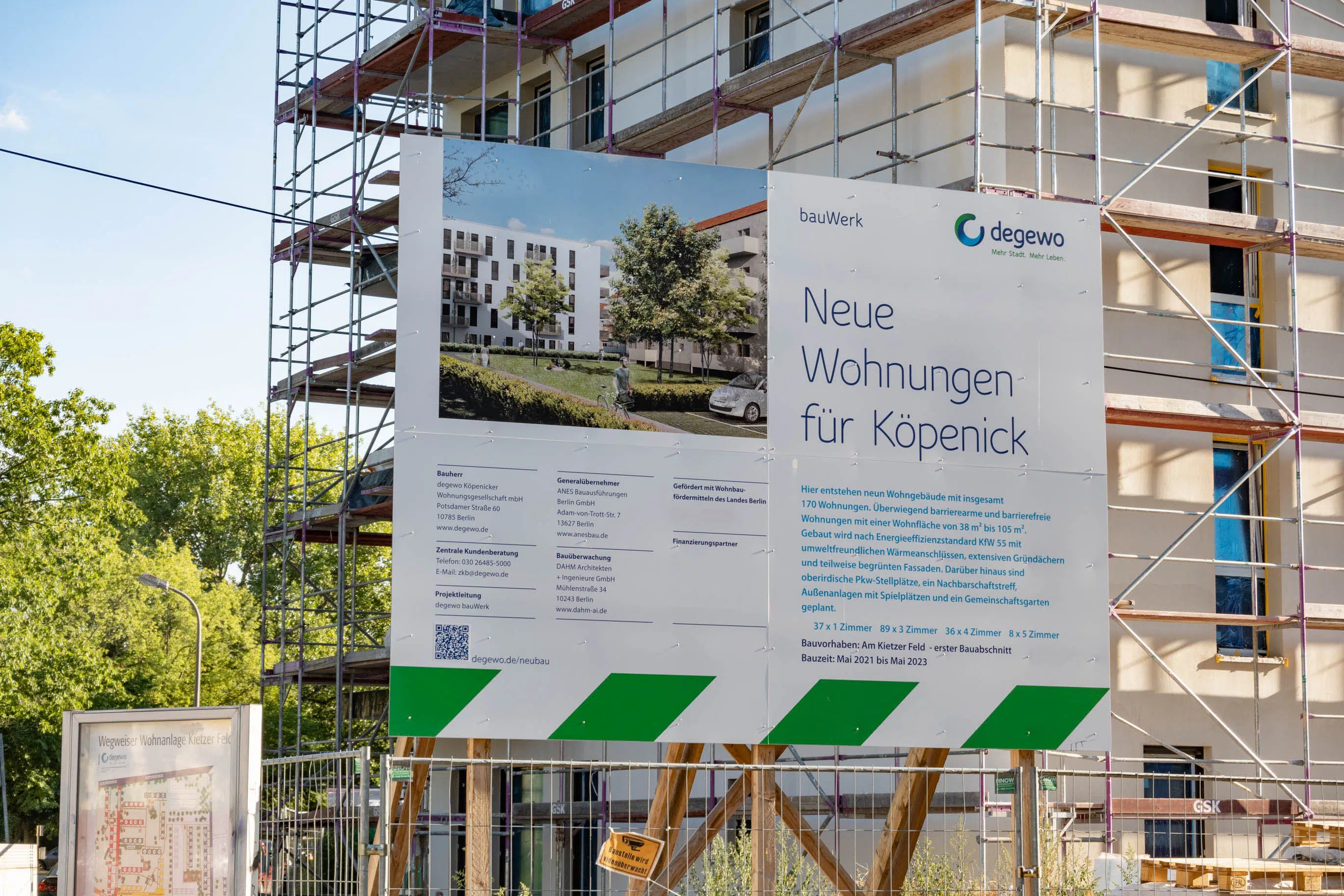 Ein Bauschild der degewo mit Informationen zu einem Wohnungsbauprojekt in Köpenick. Das Schild zeigt eine Visualisierung des fertigen Gebäudes sowie Projektdetails. Im Hintergrund ist das im Bau befindliche Gebäude mit Gerüst zu sehen.