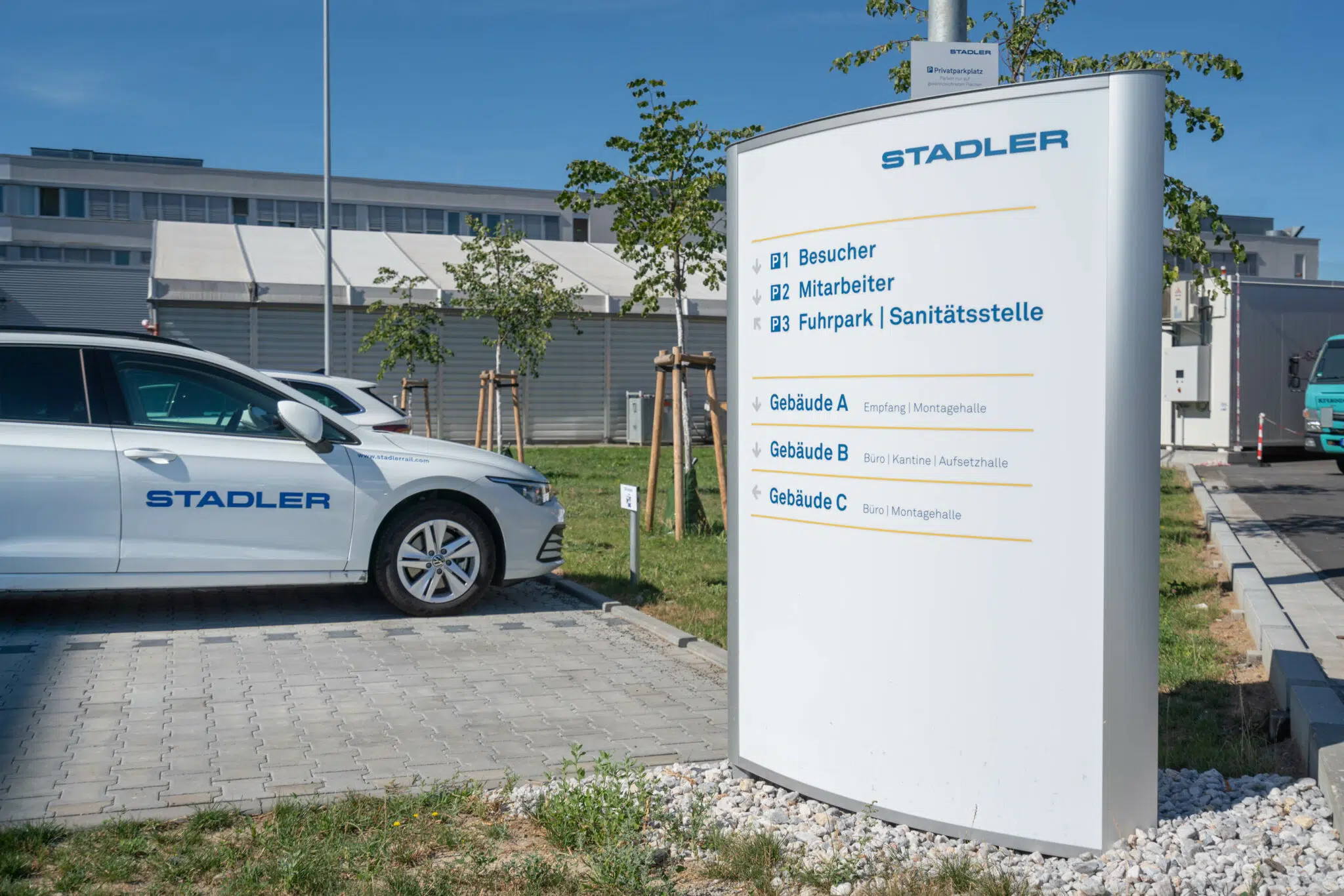 Ein Parkplatz mit einem weißen Firmenwagen mit 'Stadler'-Logo im Vordergrund. Rechts daneben eine große weiße Wegweiser-Tafel mit blauer und gelber Beschriftung, die zu Besucherparkplätzen, Mitarbeiterbereichen und verschiedenen Gebäuden auf dem Gelände führt.
