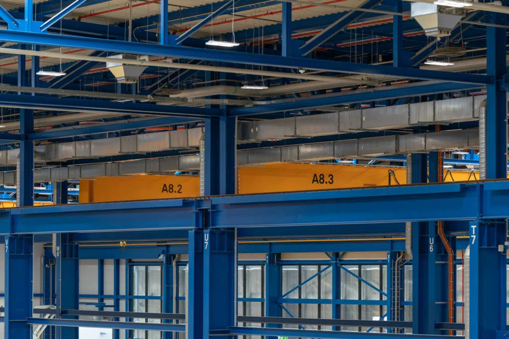 Eine Industriehalle mit blauen Stahlträgern und zwei gelben Krananlagen, die mit den Bezeichnungen 'A8.2' und 'A8.3' beschriftet sind. Über den Kränen verlaufen silberne Lüftungskanäle. Im Hintergrund sind Fenster und weitere Hallenelemente zu sehen.