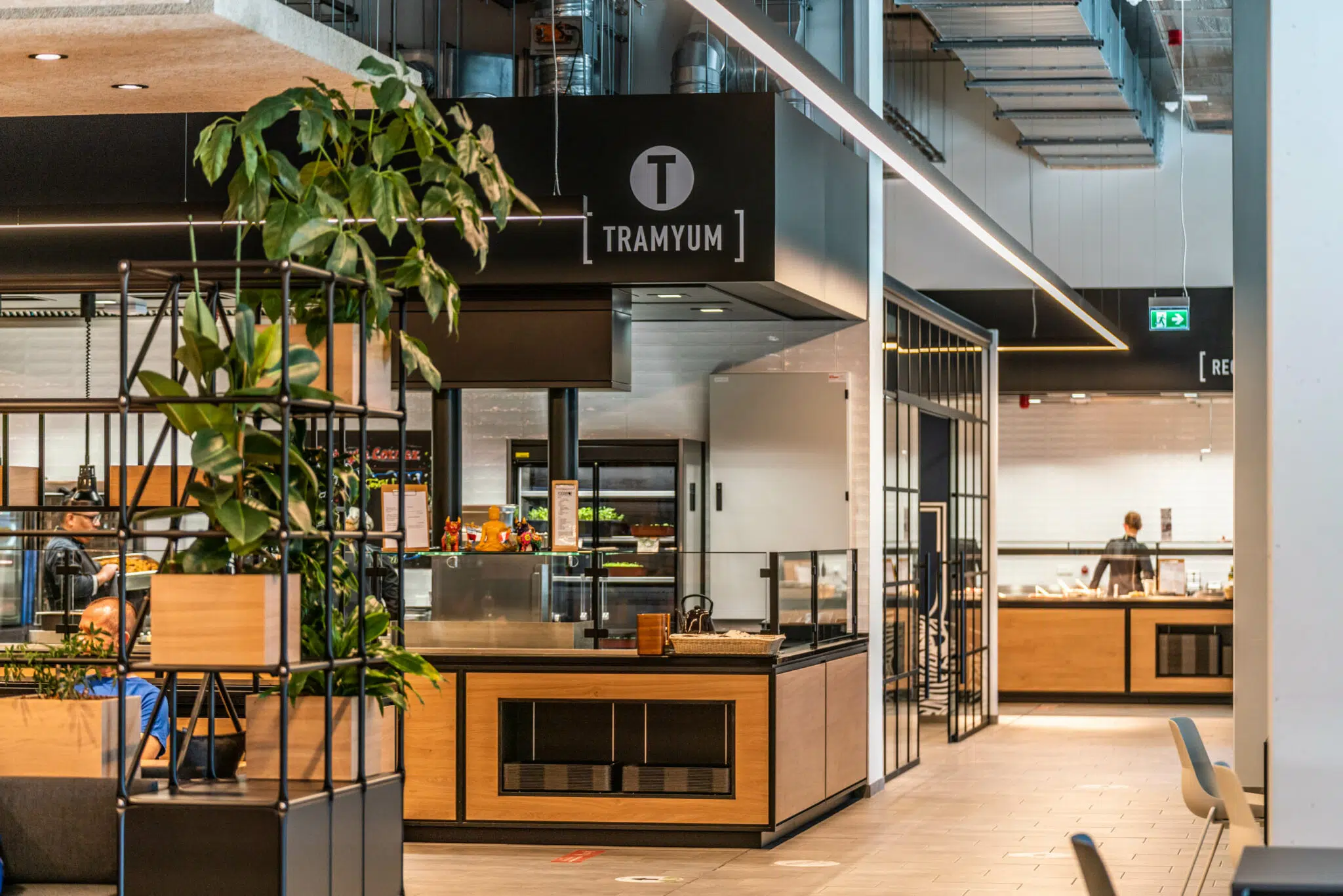 Moderne Gastronomiefläche mit einem Essensstand namens '[TRAMYUM]'. Der Bereich ist mit Holz- und Metallelementen gestaltet. Ein Regal mit Pflanzen trennt den Sitzbereich ab. Im Hintergrund sind eine offene Küche und weitere gastronomische Stände zu sehen.