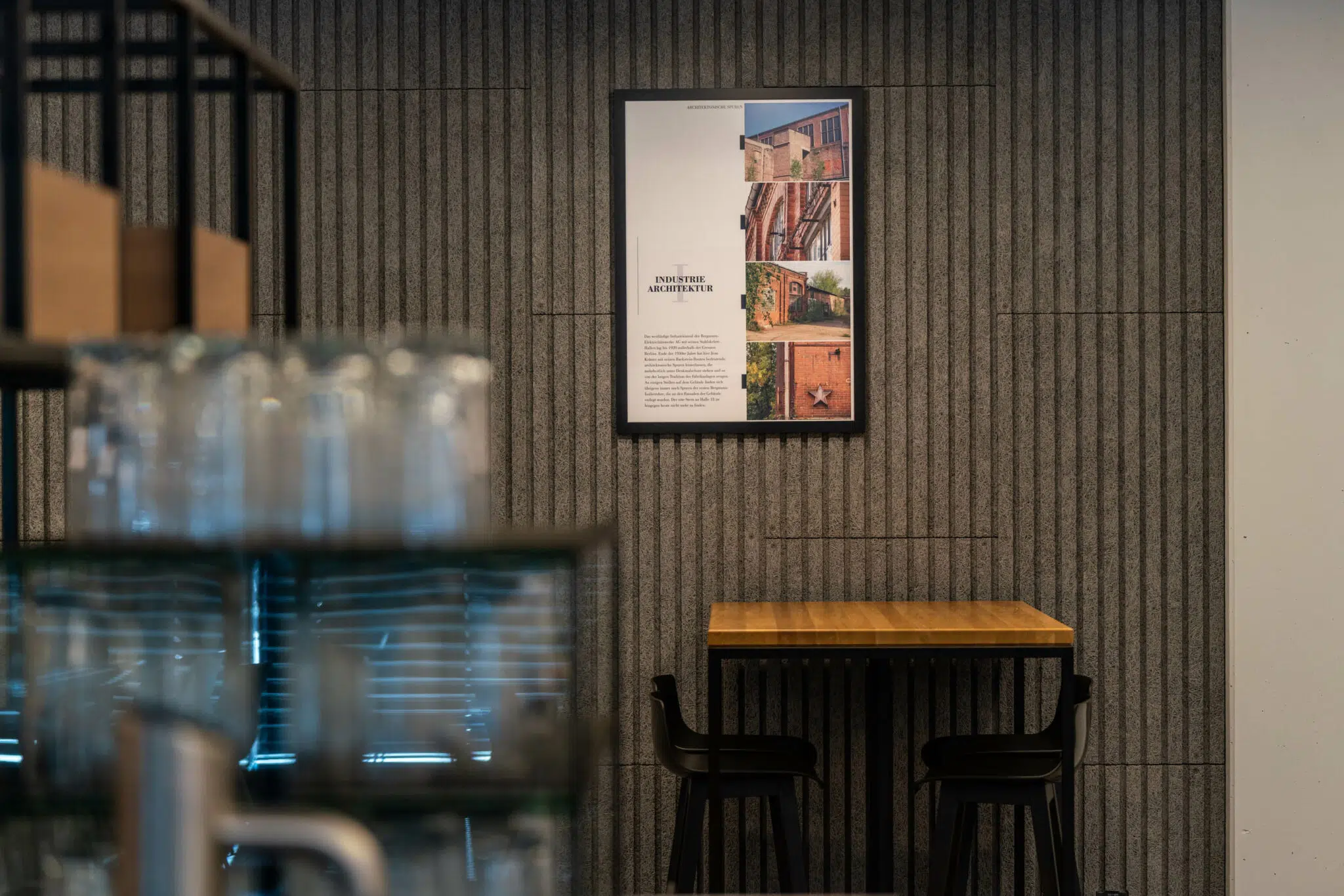 Ein modern eingerichteter Innenraum mit einer strukturierten grauen Wand, an der ein gerahmtes Poster mit Bildern von Industriearchitektur hängt. Davor steht ein schlichter Holztisch mit zwei schwarzen Stühlen. Unscharf im Vordergrund sind Gläser und ein Regal zu sehen.