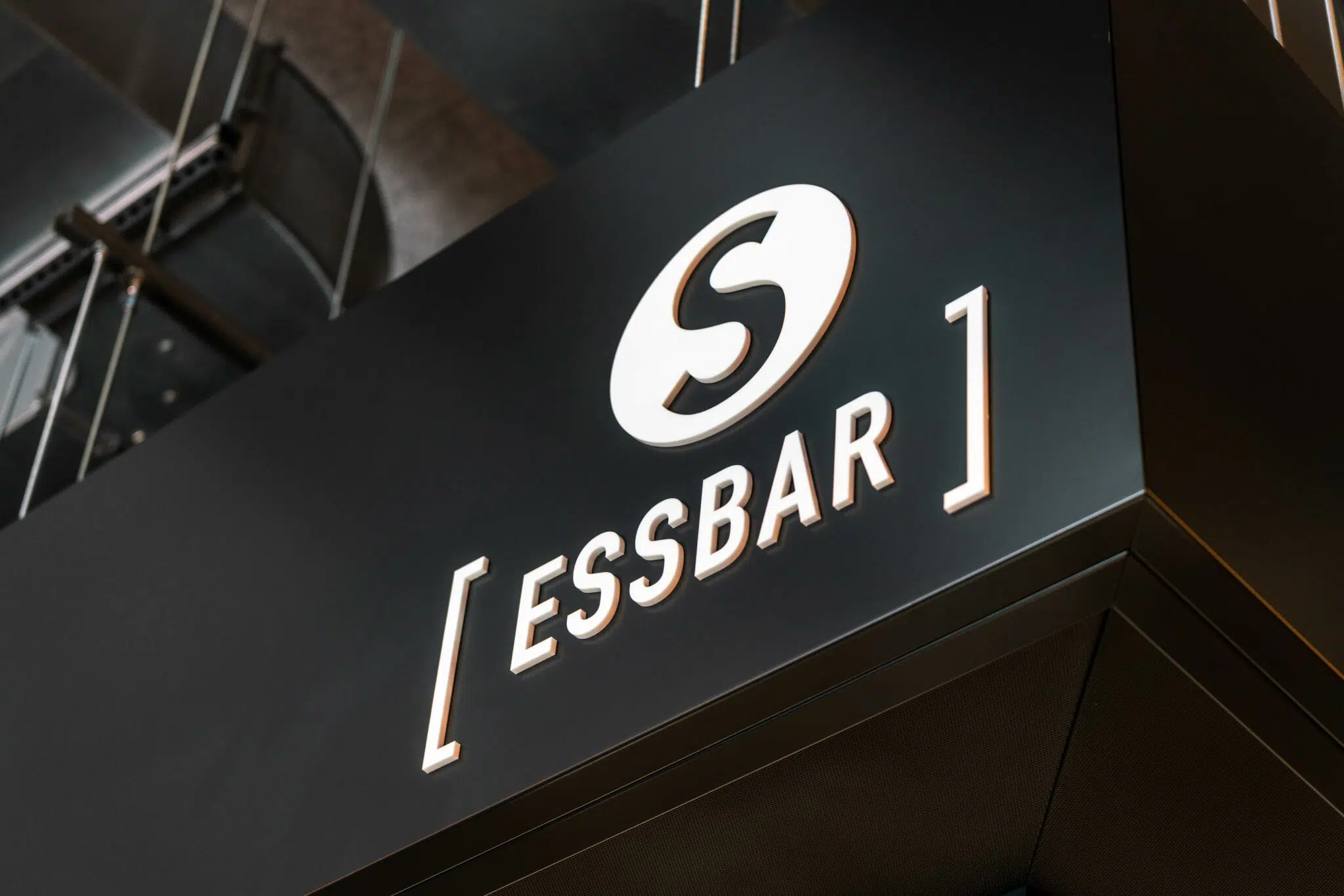 Schwarzes, modernes Schild mit weiß hinterleuchtetem Logo und Schriftzug '[ESSBAR]'. Das Logo besteht aus einem stilisierten 'S' in einem Kreis. Das Schild ist an einer Deckenkonstruktion mit Stahlseilen befestigt, im Hintergrund sind Metallstrukturen zu erkennen.