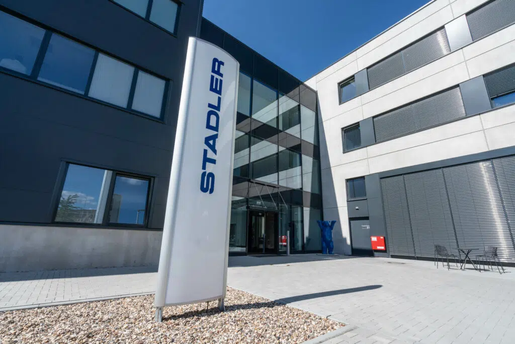 Moderner Bürokomplex mit Glasfassade und grauen Elementen. Im Vordergrund steht eine weiße Stele mit dem Schriftzug 'STADLER'. Vor dem Eingang befindet sich eine blaue Bärenstatue. Rechts sind Sitzgelegenheiten und ein roter Briefkasten an der Wand zu sehen.