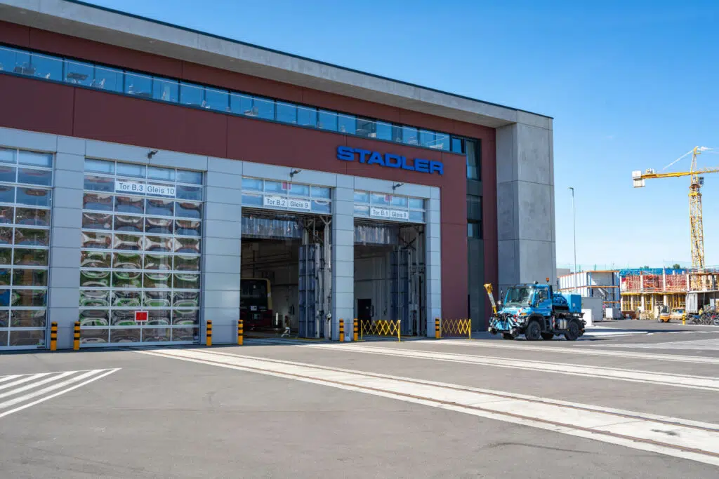 Industriegebäude mit 'STADLER'-Beschriftung. Mehrere große Tore mit Gleisnummern führen in die Halle, in der eine Bahn sichtbar ist. Ein blaues Wartungsfahrzeug steht vor dem Gebäude. Rechts im Hintergrund sind Baukräne und eine Baustelle zu sehen.