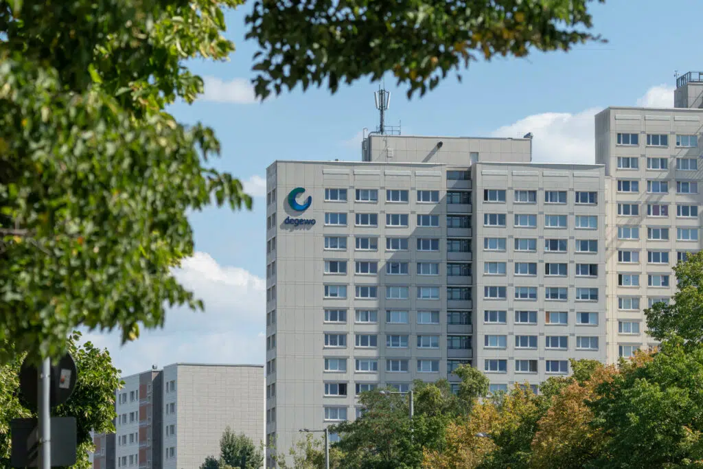 Ein großes, mehrstöckiges Wohngebäude der degewo mit zahlreichen Fenstern und einem hellgrauen Fassadendesign. Das degewo-Logo ist auf der Gebäudeseite zu sehen. Im Vordergrund rahmen grüne Baumkronen das Bild ein, während der Himmel teils bewölkt ist.
