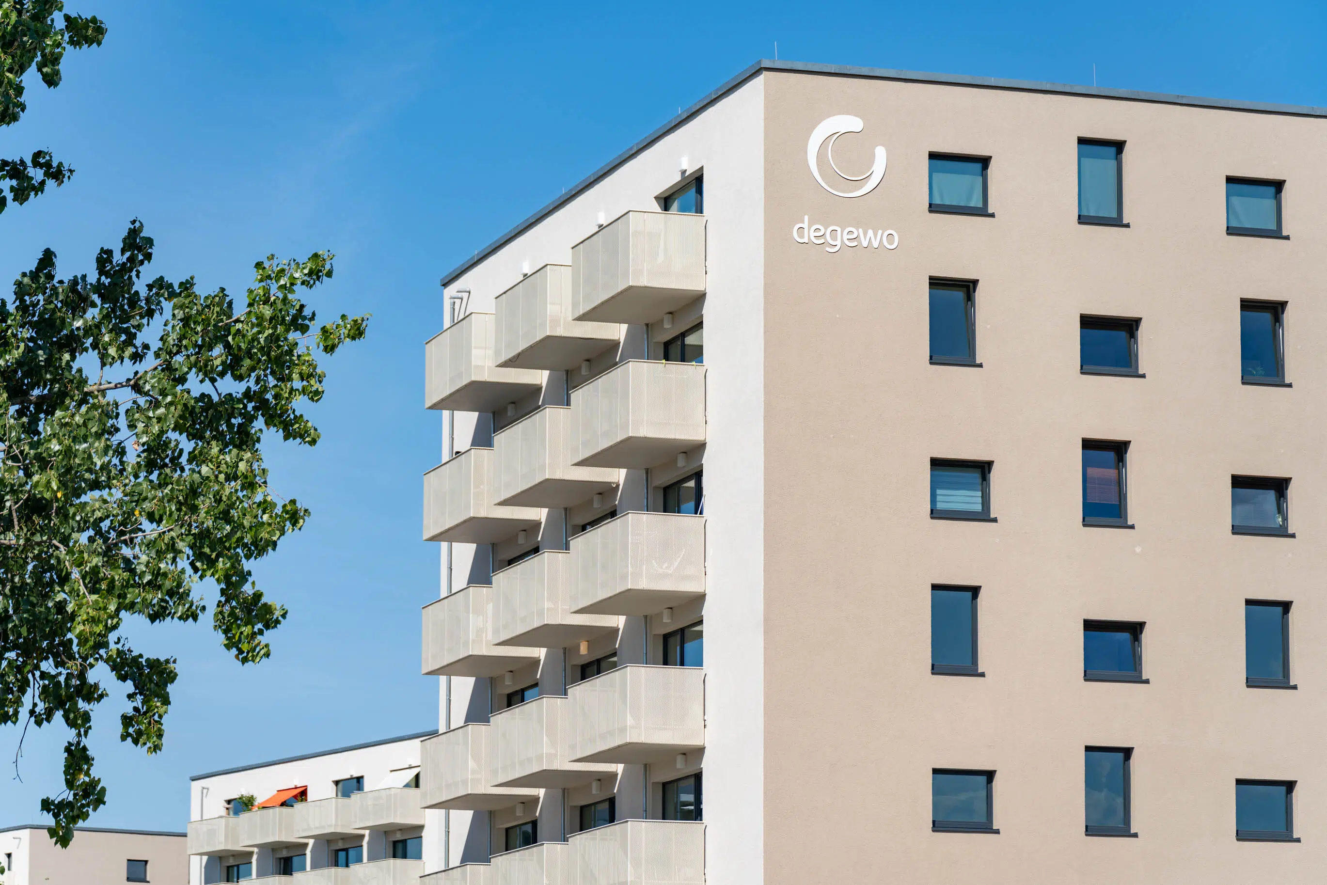 Ein modernes Wohngebäude der degewo mit mehreren Stockwerken. Die Fassade ist beige, mit dunklen Fensterrahmen und einer Reihe von hellen Balkonen. Das Logo von degewo ist an der Gebäudeseite angebracht. Links im Bild ist ein Baum mit grünen Blättern zu sehen.