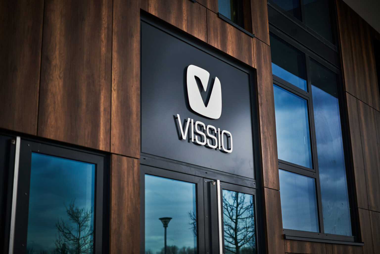 Eine moderne Gebäudefassade mit Holzoptik zeigt ein hochwertiges, dreidimensionales Firmenschild mit dem Schriftzug „VISSIO“. Die eleganten silbernen Buchstaben und das Logo heben sich klar von der schwarzen Oberfläche ab.