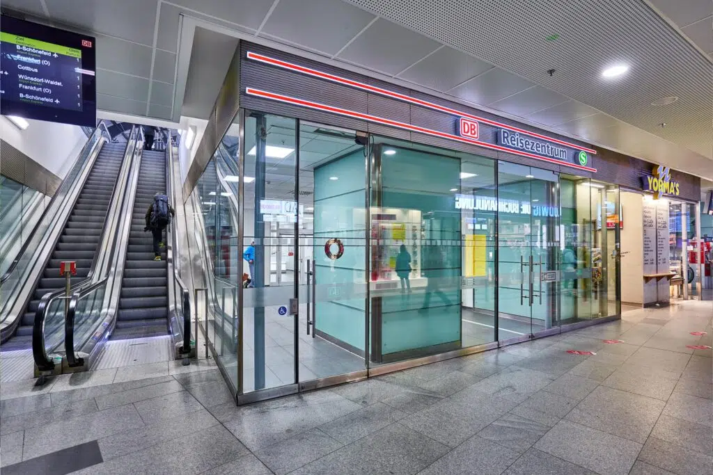 "Ein DB Reisezentrum in einem Bahnhof mit einer Glasfassade und automatischen Schiebetüren. Links eine Rolltreppe, auf der eine Person mit Rucksack nach oben fährt. Rechts ein Yorma’s-Imbiss. Spiegelungen von Reisenden sind auf den Glasscheiben sichtbar.