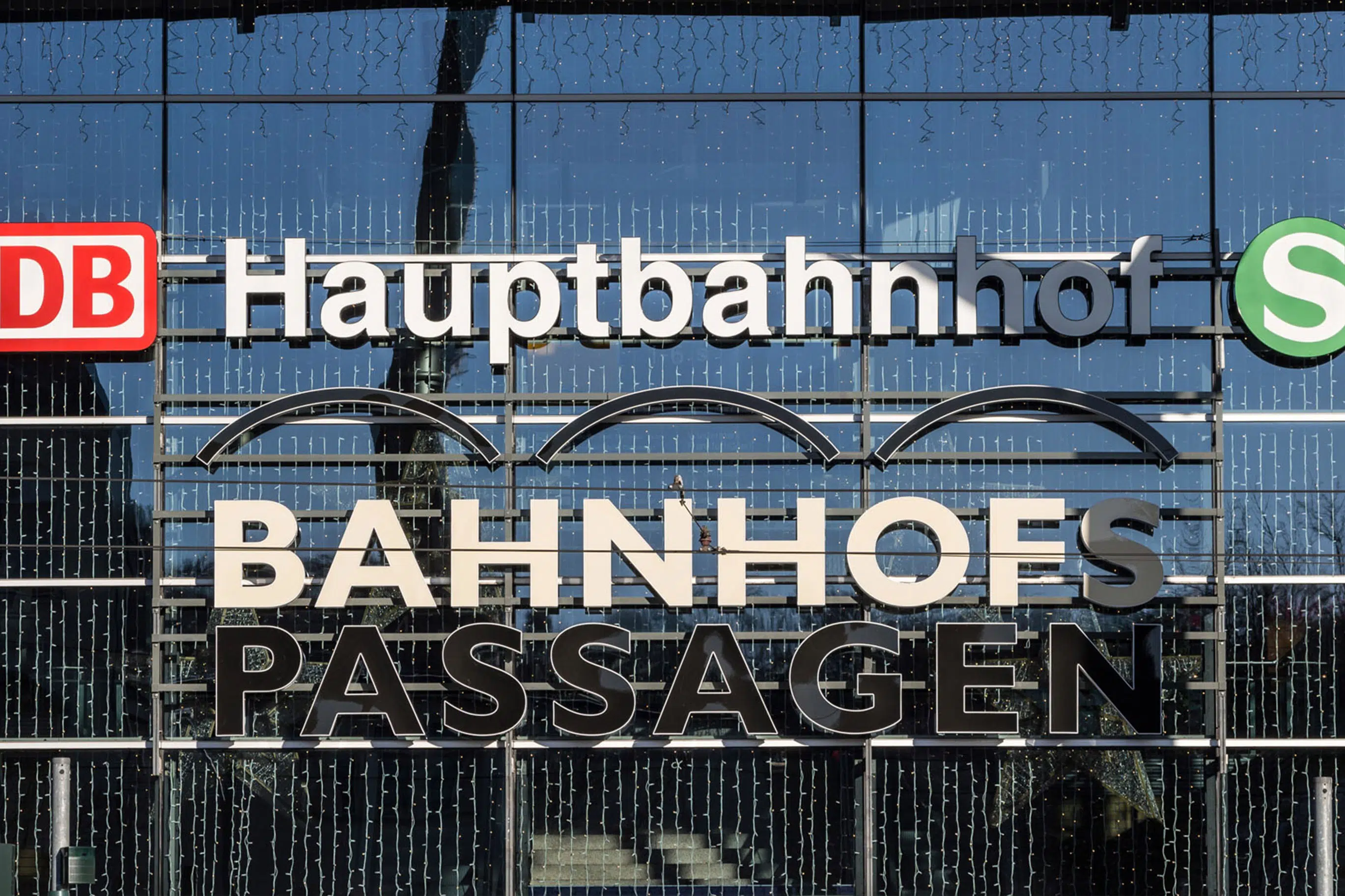 Die Glasfassade eines Hauptbahnhofs mit den Schriftzügen 'Hauptbahnhof' und 'BAHNHOFS PASSAGEN'. Logos der Deutschen Bahn (DB) und der S-Bahn sind sichtbar. Lichterketten hängen an der Fassade, und Spiegelungen der Umgebung sind im Glas zu sehen.