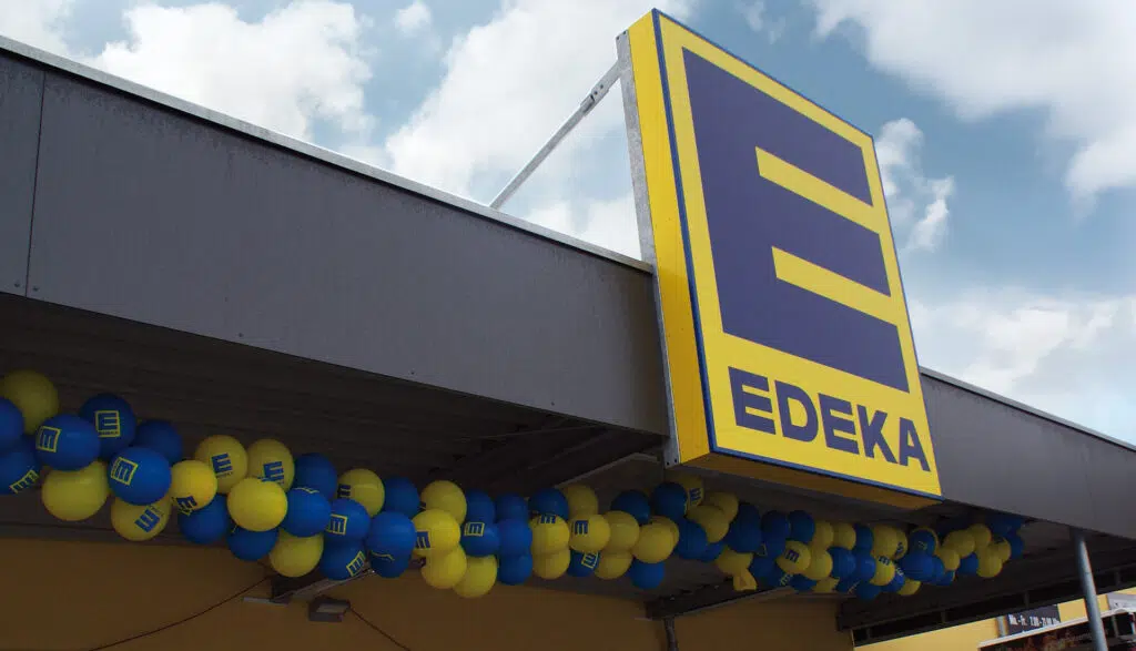Außenansicht einer EDEKA-Filiale mit großem Logo in blau-gelber Farbgebung und Dekoration aus farblich passenden Luftballons. Der Himmel im Hintergrund ist bewölkt, was der Szene ein lebendiges Ambiente verleiht.