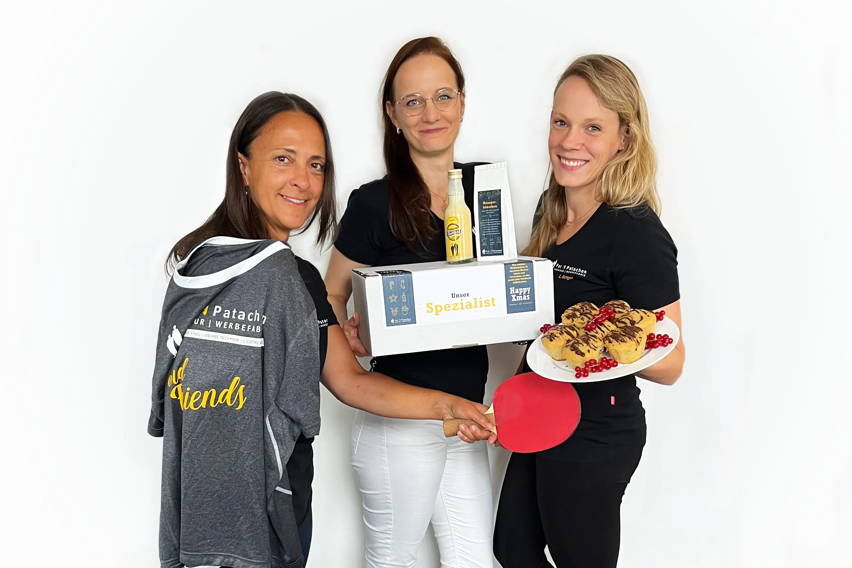 Drei gut gelaunte Frauen präsentieren kreative Geschenke und süße Leckereien. Mit Teamgeist und Freude setzen sie Akzente für eine besondere Wertschätzung. Das Branding von Pat & Patachon unterstreicht die Verbindung aus Werbetechnik und Gemeinschaft.