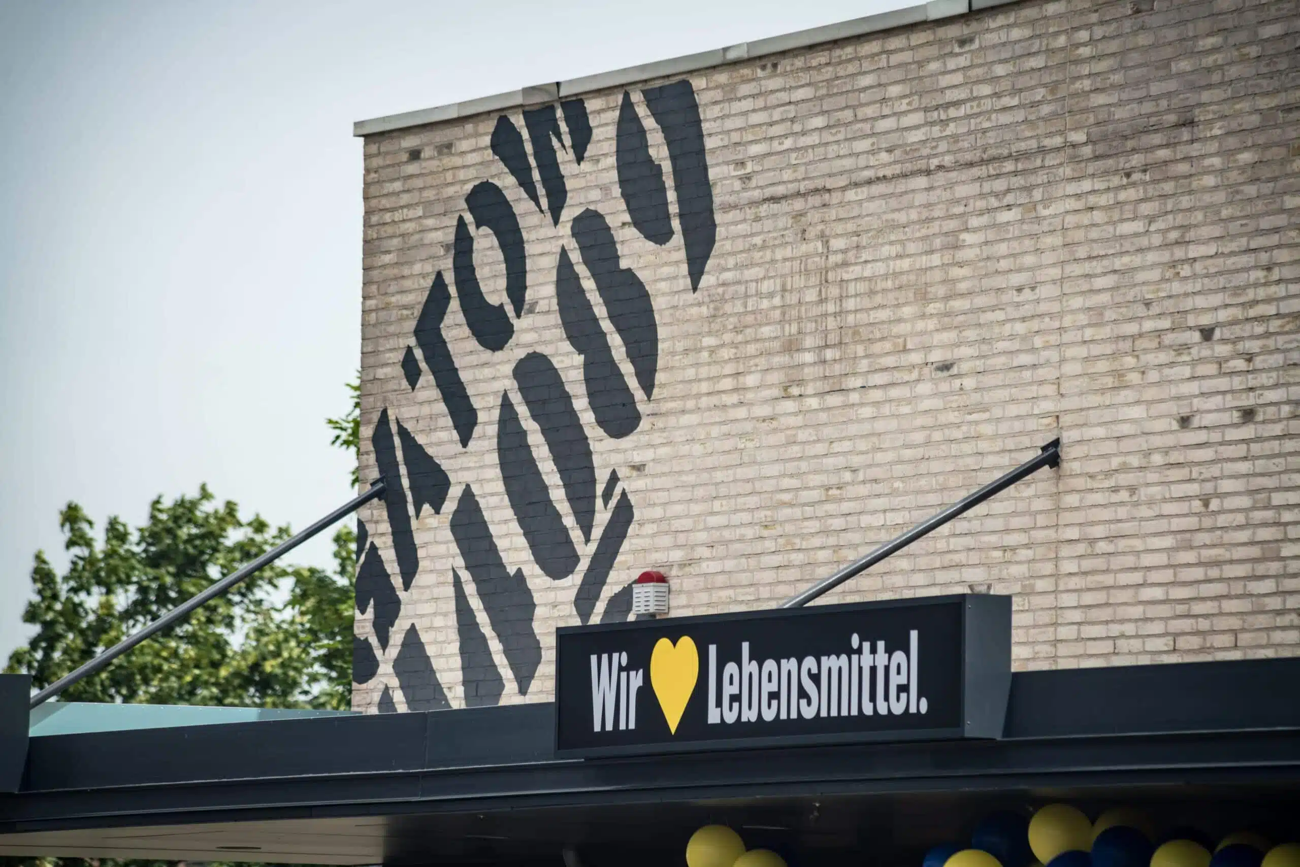 Ein EDEKA-Markt mit dem Slogan „Wir [HERZ] Lebensmittel.“ auf einem schwarzen Schild über dem Eingang. Die Backsteinwand trägt eine große, schablonenartige Aufschrift. Darunter sind gelb-blaue Ballons zur Dekoration angebracht. Eine moderne, urbane Gestaltung mit Vintage-Elementen.