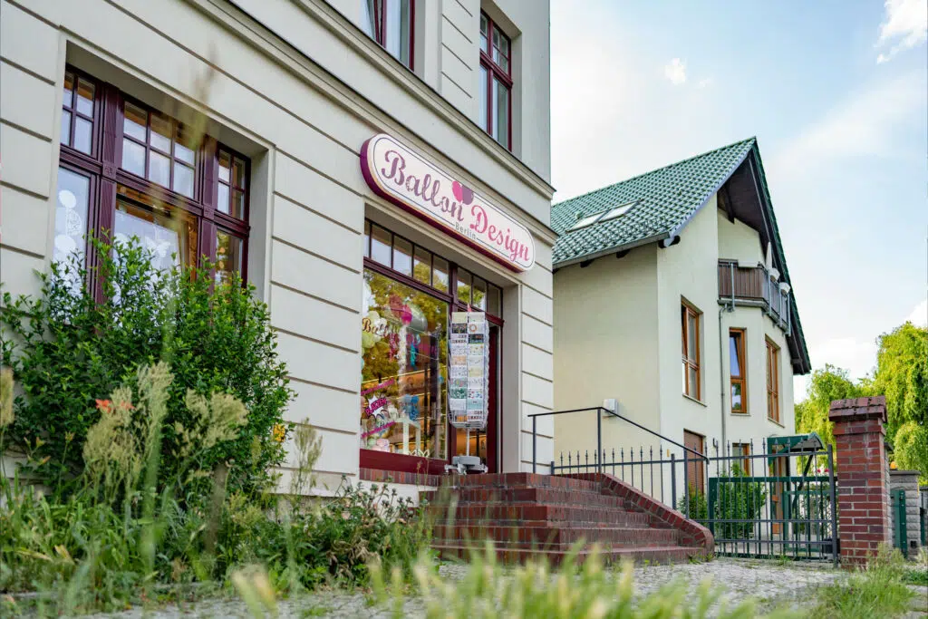 Ein Geschäft mit der Aufschrift 'Ballon Design Berlin' an der Fassade eines hellen Gebäudes mit roten Fenstern. Der Eingang ist von Ballons und einem Zeitungsständer gesäumt. Im Vordergrund wächst Gras, im Hintergrund steht ein Haus mit grünem Dach.