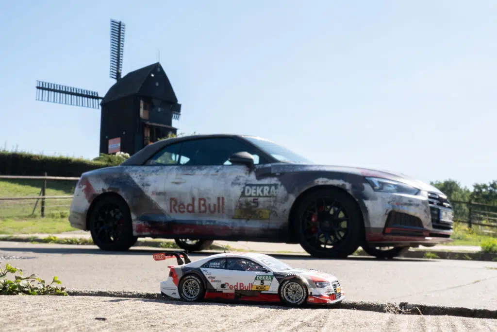 Ein im Motorsport-Look foliertes Auto mit Red Bull Branding steht auf einer Straße vor einer traditionellen Windmühle. Im Vordergrund befindet sich ein ferngesteuertes Modellauto im gleichen Design, was eine beeindruckende Perspektive und einen kreativen Größenvergleich schafft.