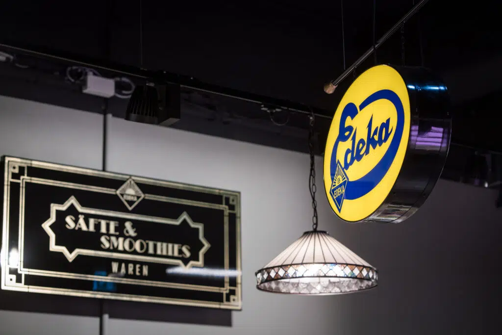 Edles Retro-Design bei EDEKA: Eine stilvolle Saft- & Smoothie-Beschilderung in Schwarz-Gold im Art-Déco-Stil kombiniert mit einer nostalgischen Tiffany-Lampe. Das beleuchtete runde EDEKA-Logo verleiht dem Ganzen eine moderne Note mit Vintage-Charme.