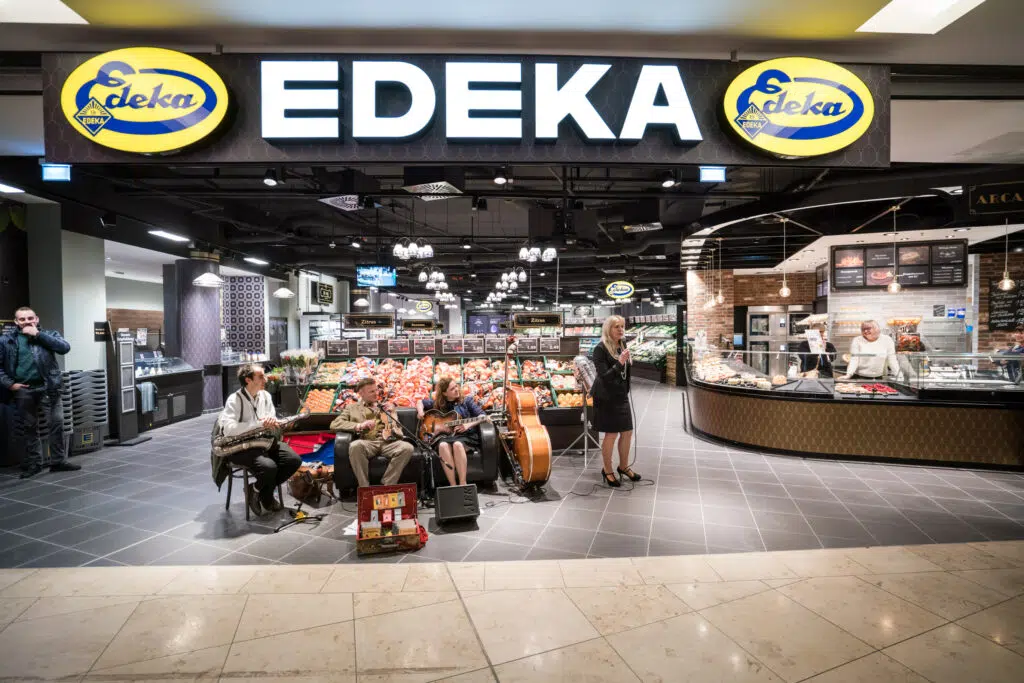 Ein stilvoller EDEKA-Markt mit beleuchtetem Logo und modernem Interieur. Im Eingangsbereich spielt eine Live-Band mit Saxophon, Kontrabass und Gitarre, während eine Sängerin performt. Frische Obst- und Gemüseabteilung im Hintergrund, daneben eine Feinkosttheke.