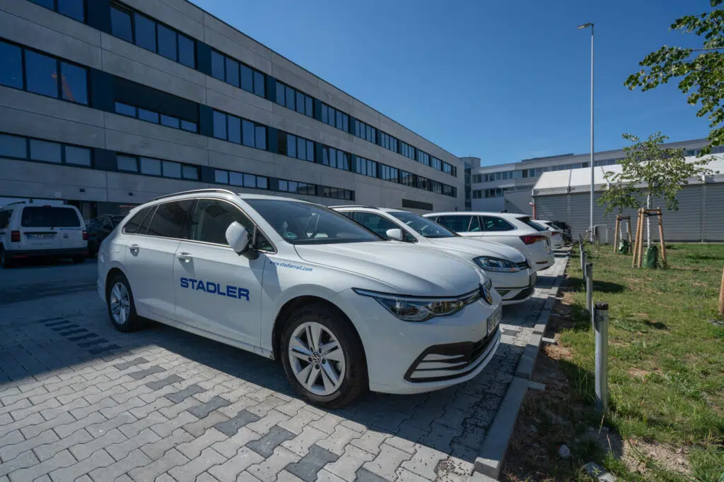 Weiße VW-Fahrzeuge mit Stadler-Branding in blauer Schrift auf einem Firmenparkplatz. Im Hintergrund moderne Bürogebäude mit großen Fenstern, ein Laternenmast und junge Bäume. Klare Schatten zeigen sonniges Wetter.