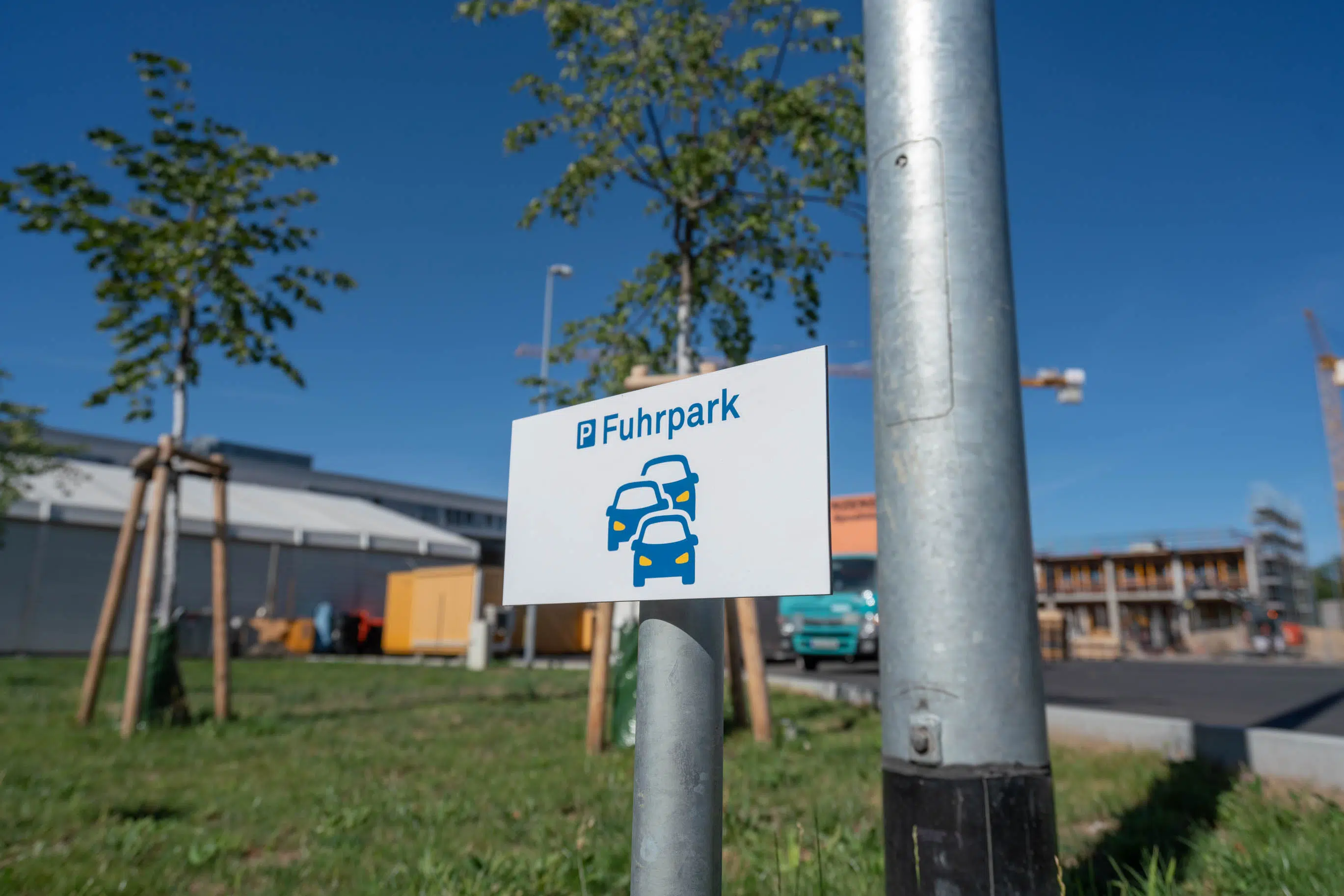 ChatGPT:Nahaufnahme eines Schildes mit der Aufschrift „P Fuhrpark“ und einer stilisierten Fahrzeuggrafik. Es ist an einem Metallpfosten befestigt. Im Hintergrund befinden sich junge Bäume, Baucontainer und ein modernes Gebäude mit Baukränen unter blauem Himmel.