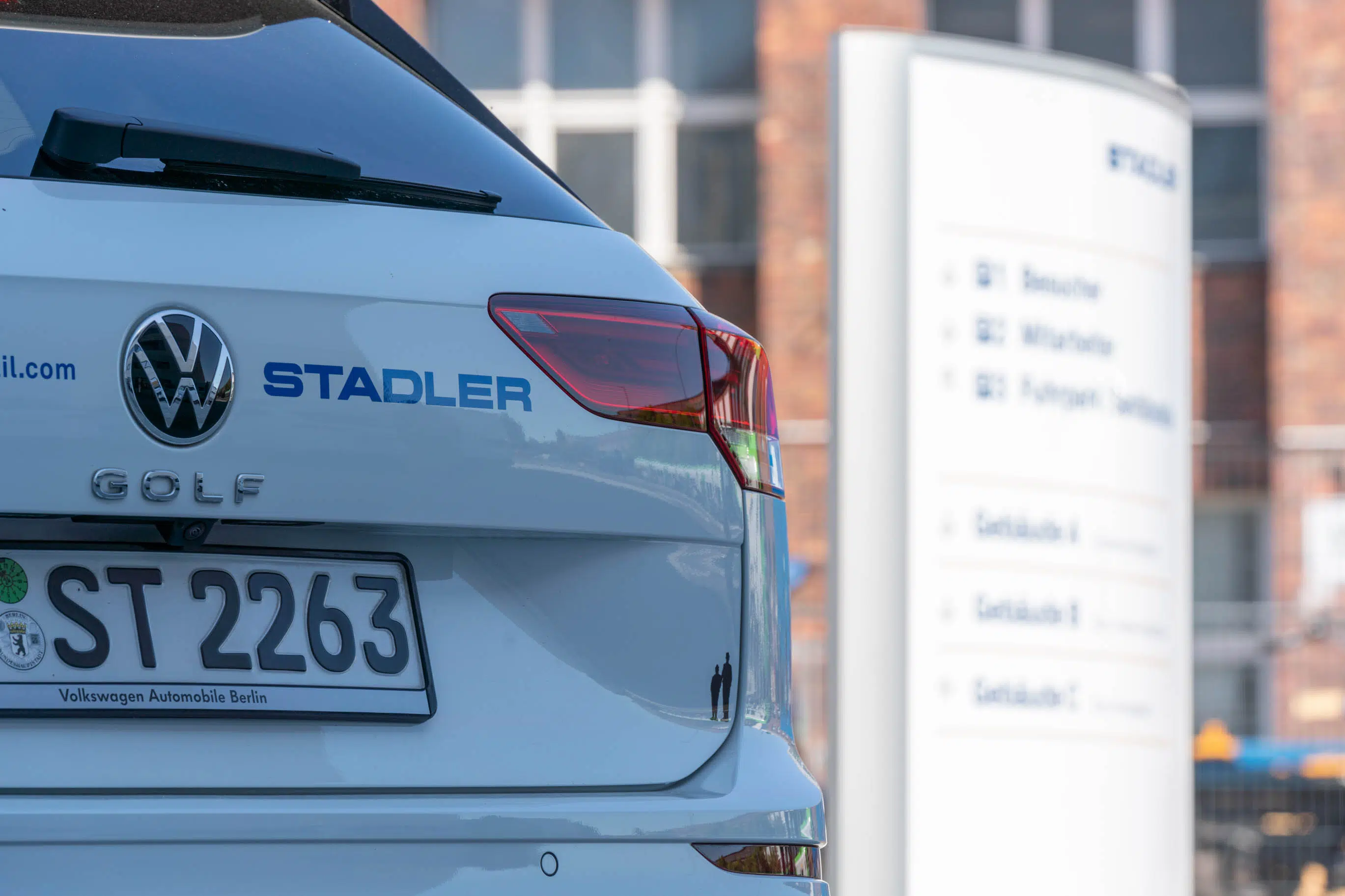 Rückansicht eines weißen VW Golf mit Stadler-Branding in blauer Schrift. Im Hintergrund eine unscharfe Wegweiser-Tafel mit Gebäudehinweisen. Das Fahrzeug trägt ein dezentes P&P Logo in der unteren Ecke.