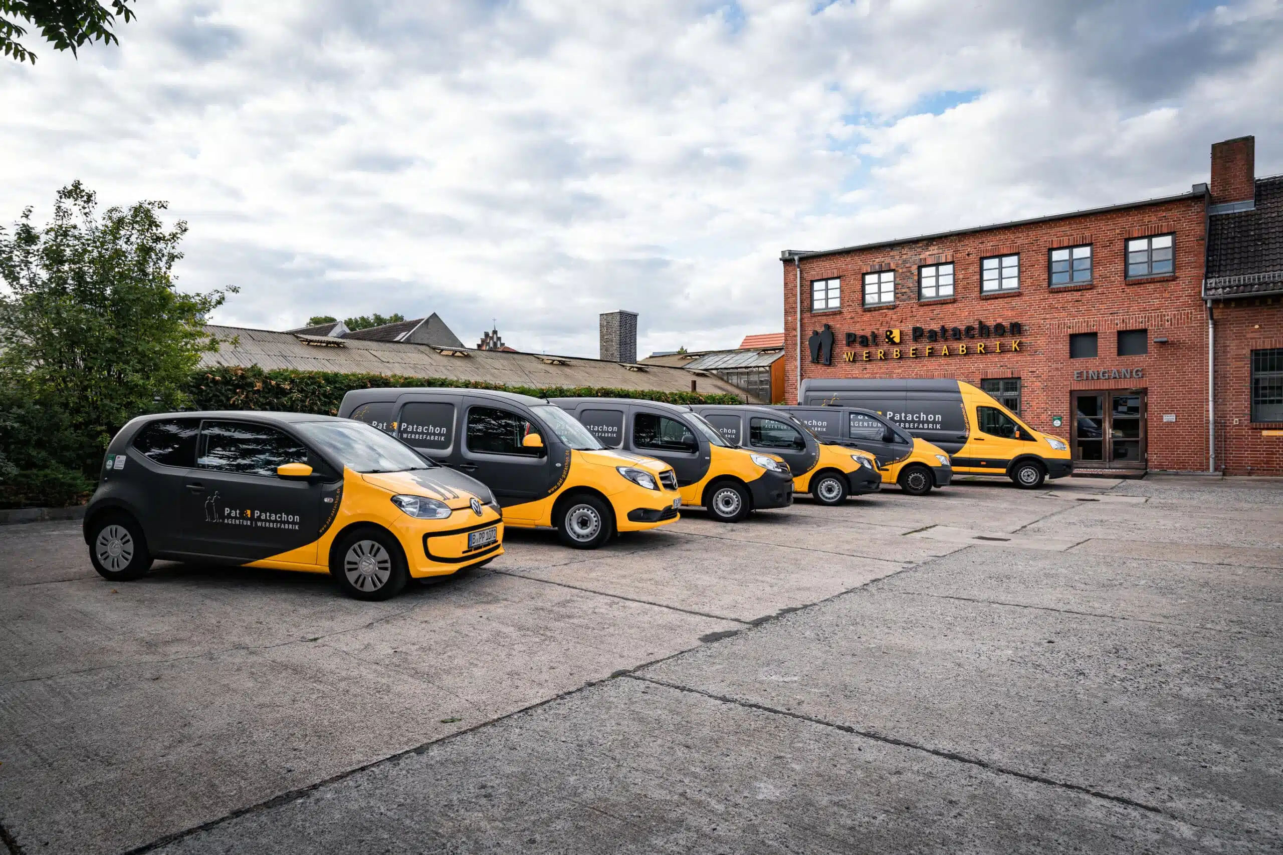 Ein Firmenfuhrpark von Pat & Patachon mit mehreren schwarz-gelb folierten Fahrzeugen, darunter ein VW Up, mehrere Mercedes Citan und ein Ford Transit. Die Fahrzeuge stehen vor einem Backsteingebäude mit dem Firmennamen „Pat & Patachon Werbefabrik“.