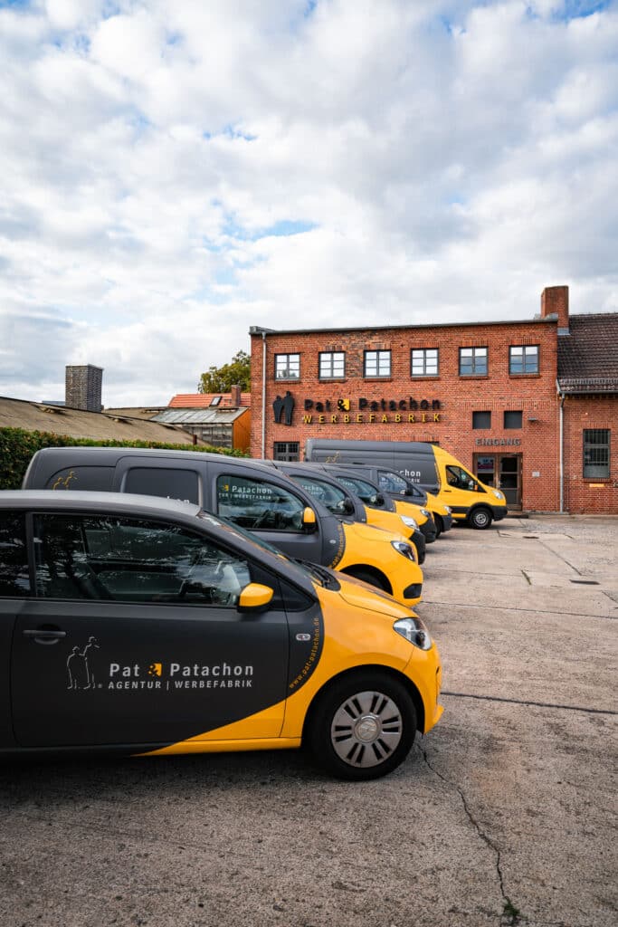 Eine Reihe von schwarz-gelben Firmenfahrzeugen mit dem Logo von "Pat & Patachon" steht vor einem Backsteingebäude mit der Aufschrift „Pat & Patachon Werbefabrik“. Der Himmel ist bewölkt, und der Hof besteht aus Beton.