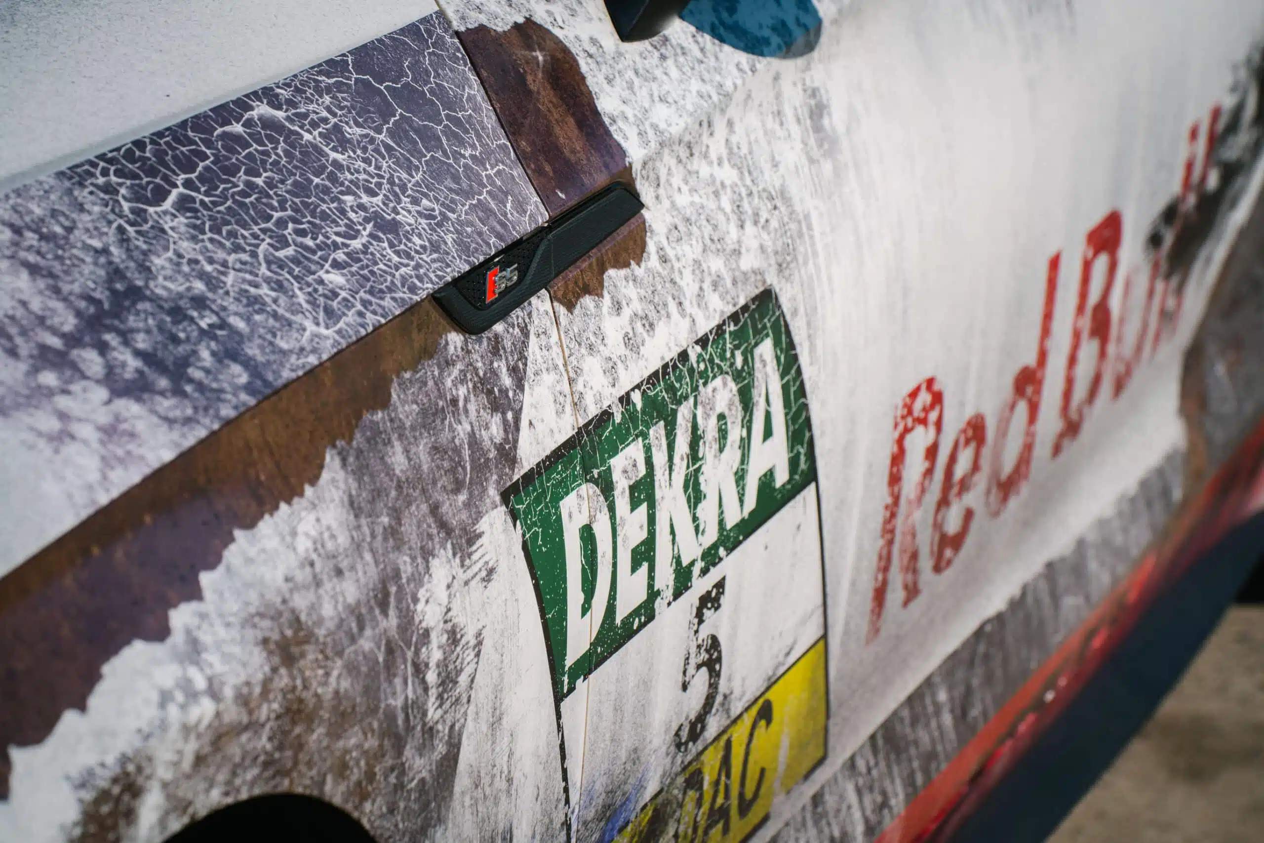 Eine Fahrzeugfolierung mit Patina-Optik zeigt realistische Rost- und Abnutzungseffekte. Auf der Tür sind Dekra- und Red Bull-Logos in verwittertem Stil zu sehen. Das Design verleiht dem Auto einen authentischen, gebrauchten Rennsport-Look.