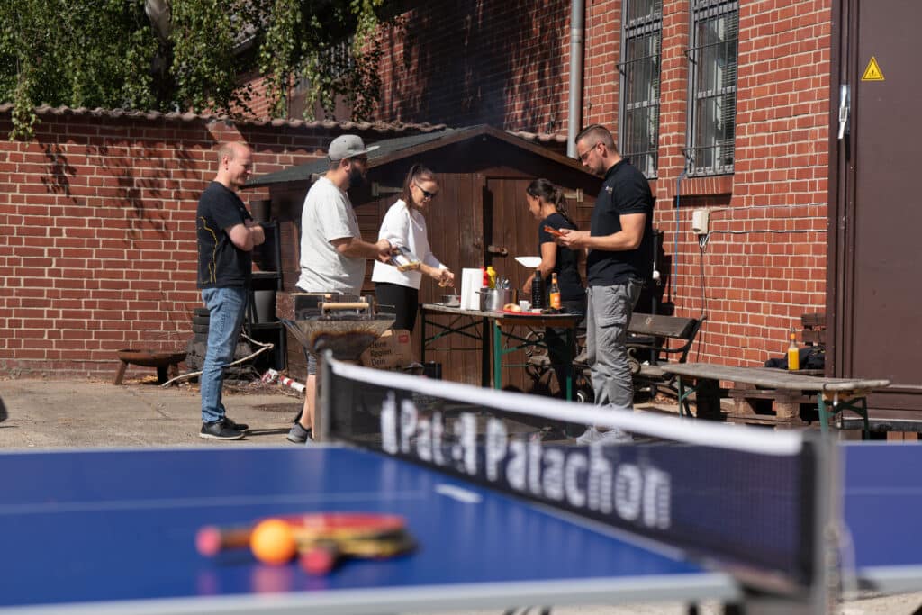 Mitarbeiter von Pat & Patachon genießen ein Grillfest auf dem Firmengelände. Im Vordergrund steht eine Tischtennisplatte mit Schlägern und Bällen, während im Hintergrund Kollegen am Grill und einem Tisch stehen, entspannt plaudern und das Essen vorbereiten.