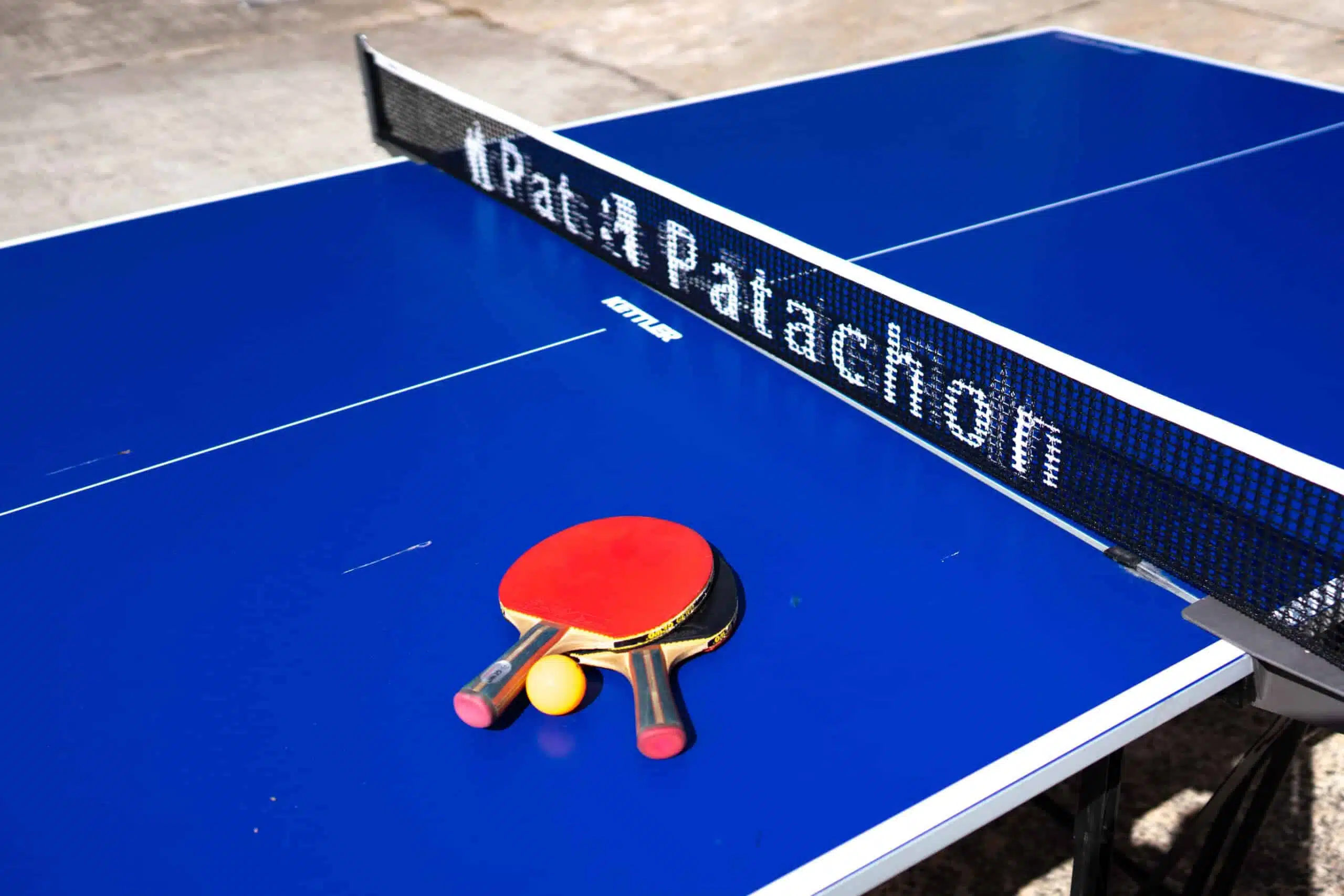 Nahaufnahme einer blauen Tischtennisplatte mit zwei Schlägern und einem gelben Ball. Das Netz trägt den Schriftzug „Pat & Patachon“. Die Szene ist im Freien, das Sonnenlicht wirft Schatten auf die Platte.