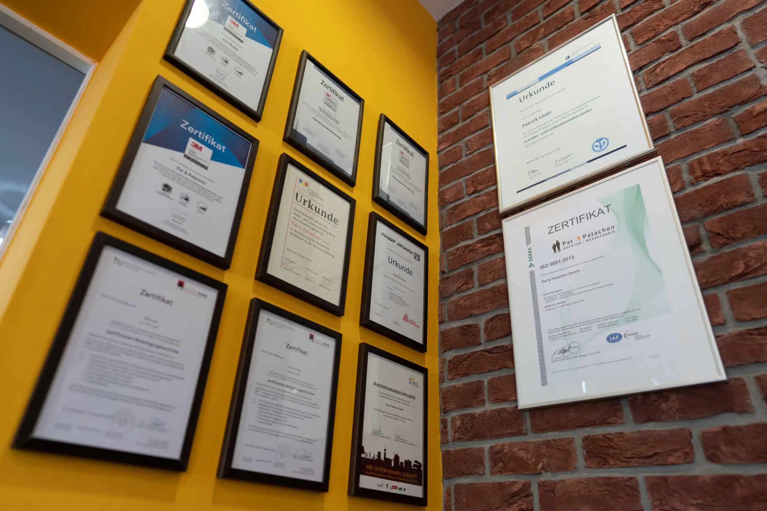 Eine Wand mit mehreren eingerahmten Zertifikaten und Urkunden. Die linke Seite ist gelb gestrichen, die rechte besteht aus Backstein. Die Auszeichnungen sind ordentlich angeordnet und zeigen verschiedene Qualifikationen und Anerkennungen von Pat & Patachon.