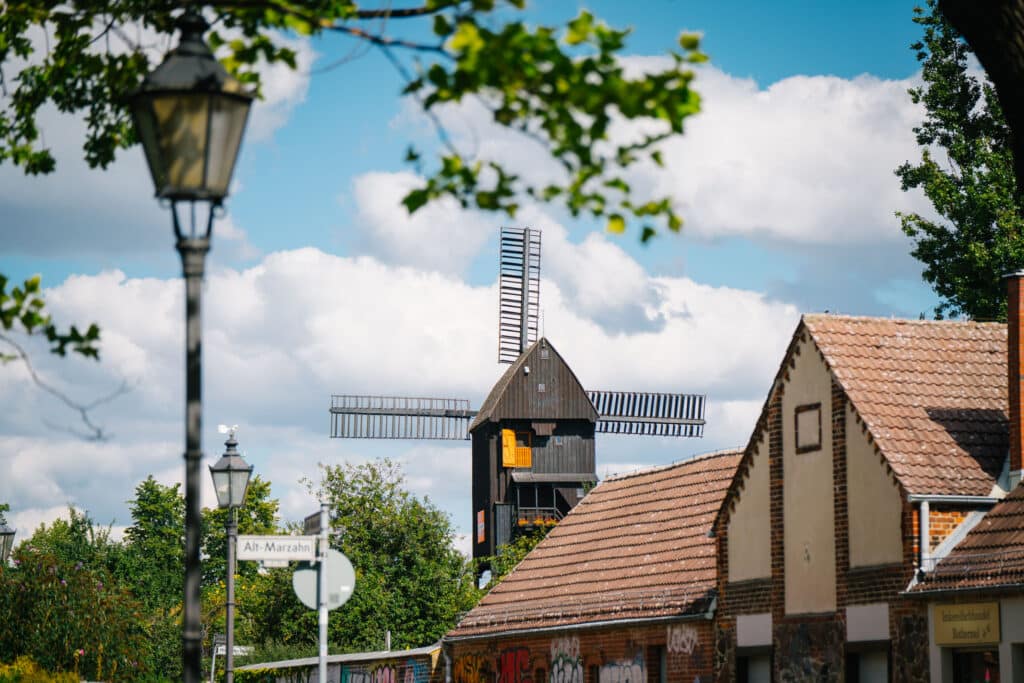 Ein schöner Blick auf eine historische Windmühle in Alt-Marzahn, Berlin. Im Vordergrund sind Laternen, Straßenschilder und traditionelle Gebäude mit Backsteinfassaden und roten Dächern zu sehen. Die Windmühle, mit ihren dunklen Holzwänden und großen Flügeln, hebt sich vor einem Himmel mit weißen Wolken und grünen Bäumen ab.