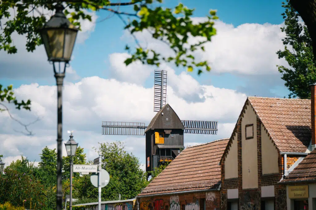 Ein schöner Blick auf eine historische Windmühle in Alt-Marzahn, Berlin. Im Vordergrund sind Laternen, Straßenschilder und traditionelle Gebäude mit Backsteinfassaden und roten Dächern zu sehen. Die Windmühle, mit ihren dunklen Holzwänden und großen Flügeln, hebt sich vor einem Himmel mit weißen Wolken und grünen Bäumen ab.