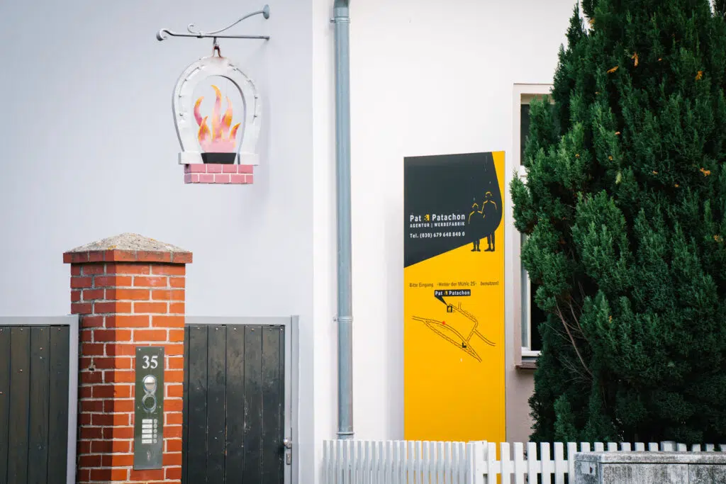 Ein Eingang mit einem Firmenschild von „Pat & Patachon Agentur | Werbefabrik“ in Gelb und Schwarz, das den Standort und eine Wegbeschreibung zeigt. Links ein Backsteinpfeiler mit Klingelanlage, darüber ein dekoratives Hufeisen mit Flammenmotiv.