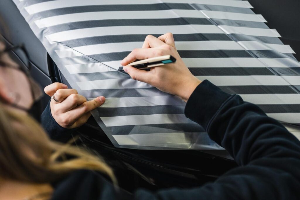 Das Bild zeigt eine Person, die mit einem Cutter präzise eine Folie mit schwarz-weißen Streifen zuschneidet und auf eine Oberfläche aufbringt. Die Person trägt eine schwarze Jacke und eine Brille. Die Folie wird mit beiden Händen gespannt und sorgfältig verarbeitet.