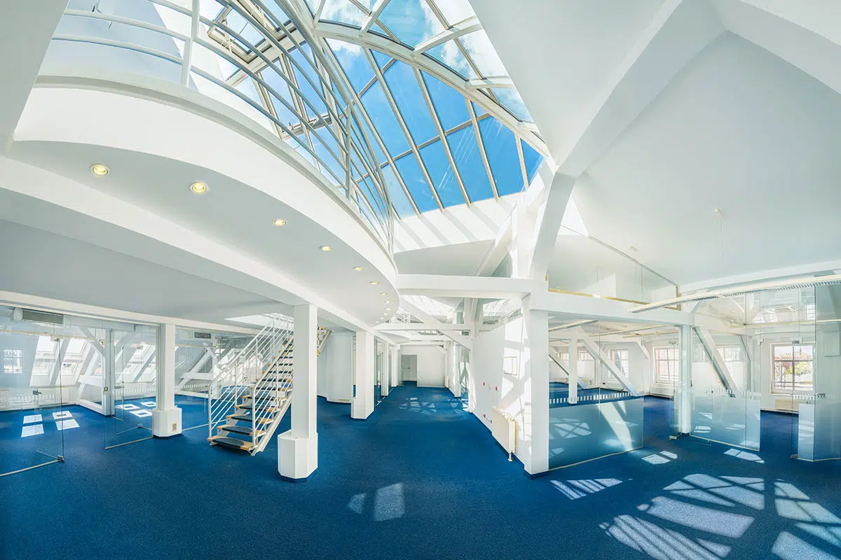 Das Bild zeigt einen modernen, lichtdurchfluteten Innenraum mit einer großen Glasdachkonstruktion. Die offene Architektur mit weißen Stahlträgern und Glaswänden schafft eine helle Atmosphäre. Eine Treppe führt auf eine erhöhte Ebene, während der Boden mit blauem Teppich ausgelegt ist.