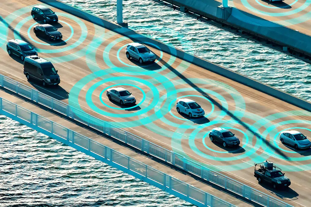 Das Bild zeigt eine Luftaufnahme einer Brücke mit mehreren fahrenden Autos. Blaue Kreise um die Fahrzeuge symbolisieren drahtlose Kommunikation oder autonome Fahrtechnologie. Im Hintergrund ist Wasser zu sehen.
