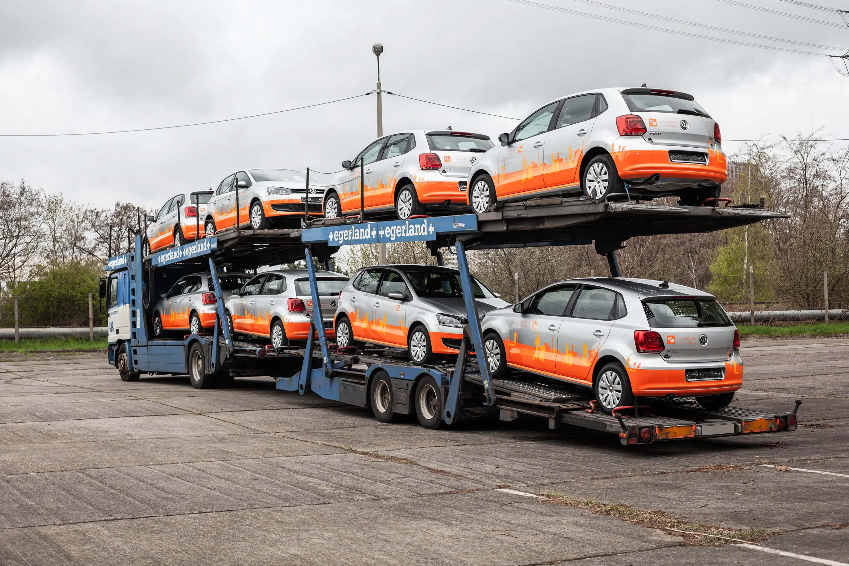 Ein Autotransporter beladen mit silbernen Volkswagen Polo, die eine auffällige orangefarbene Fahrzeugfolierung mit Stadtsilhouette tragen. Die Fahrzeuge sind für den Transport gesichert, der Himmel ist bewölkt.