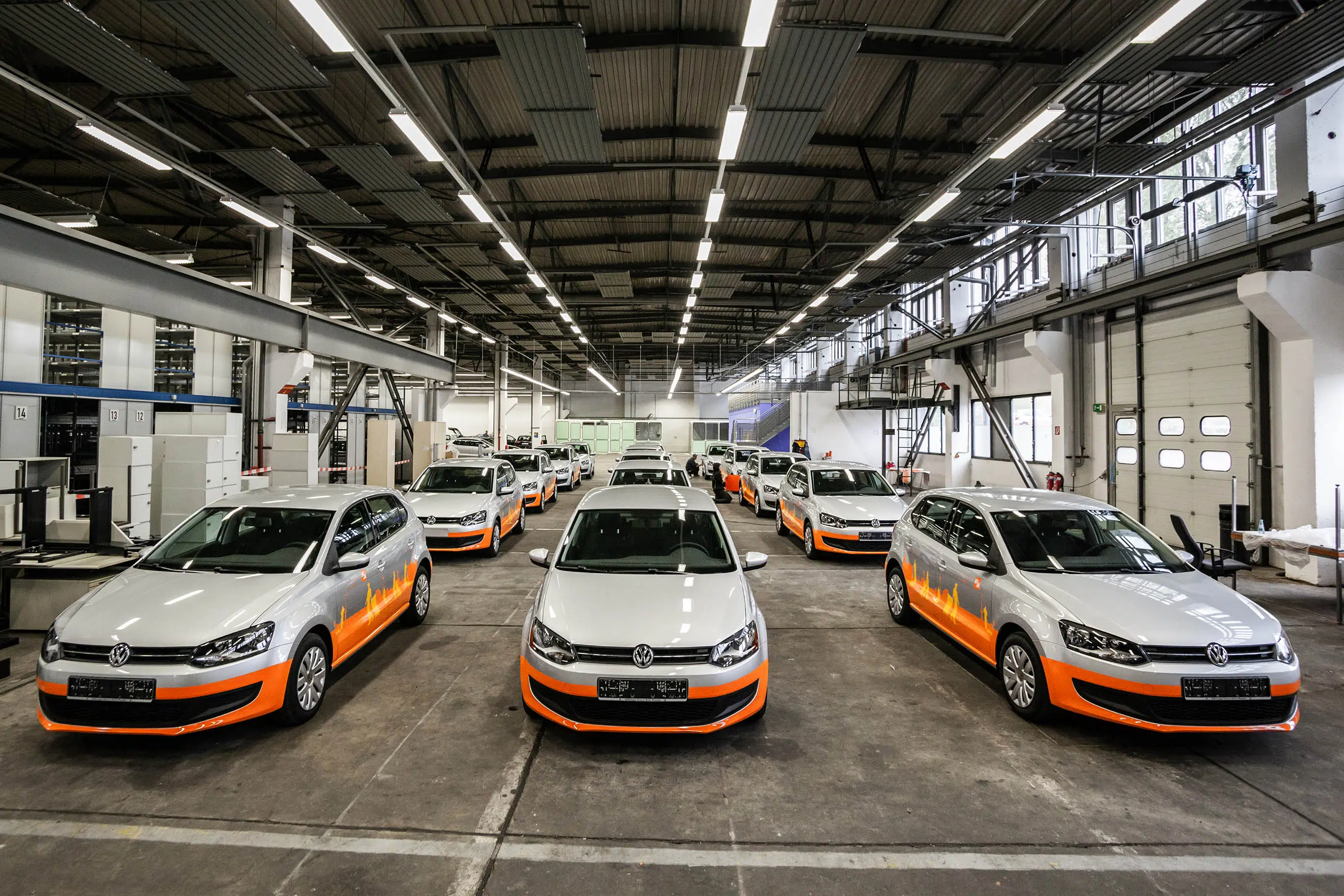Mehrere silberne Volkswagen Polo mit orangefarbener Folierung stehen in einer großen, gut beleuchteten Werkshalle in Reihen angeordnet. Die Fahrzeuge sind fertig foliert und bereit für den Einsatz.