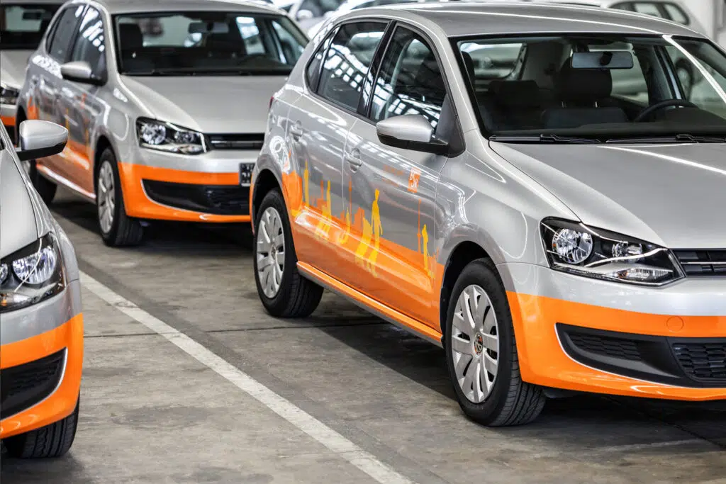 Mehrere silberne Volkswagen Polo mit auffälliger Fahrzeugfolierung in Orange und einer stilisierten Stadtsilhouette stehen in einer Tiefgarage. Die reflektierenden Scheiben spiegeln die Deckenbeleuchtung wider.