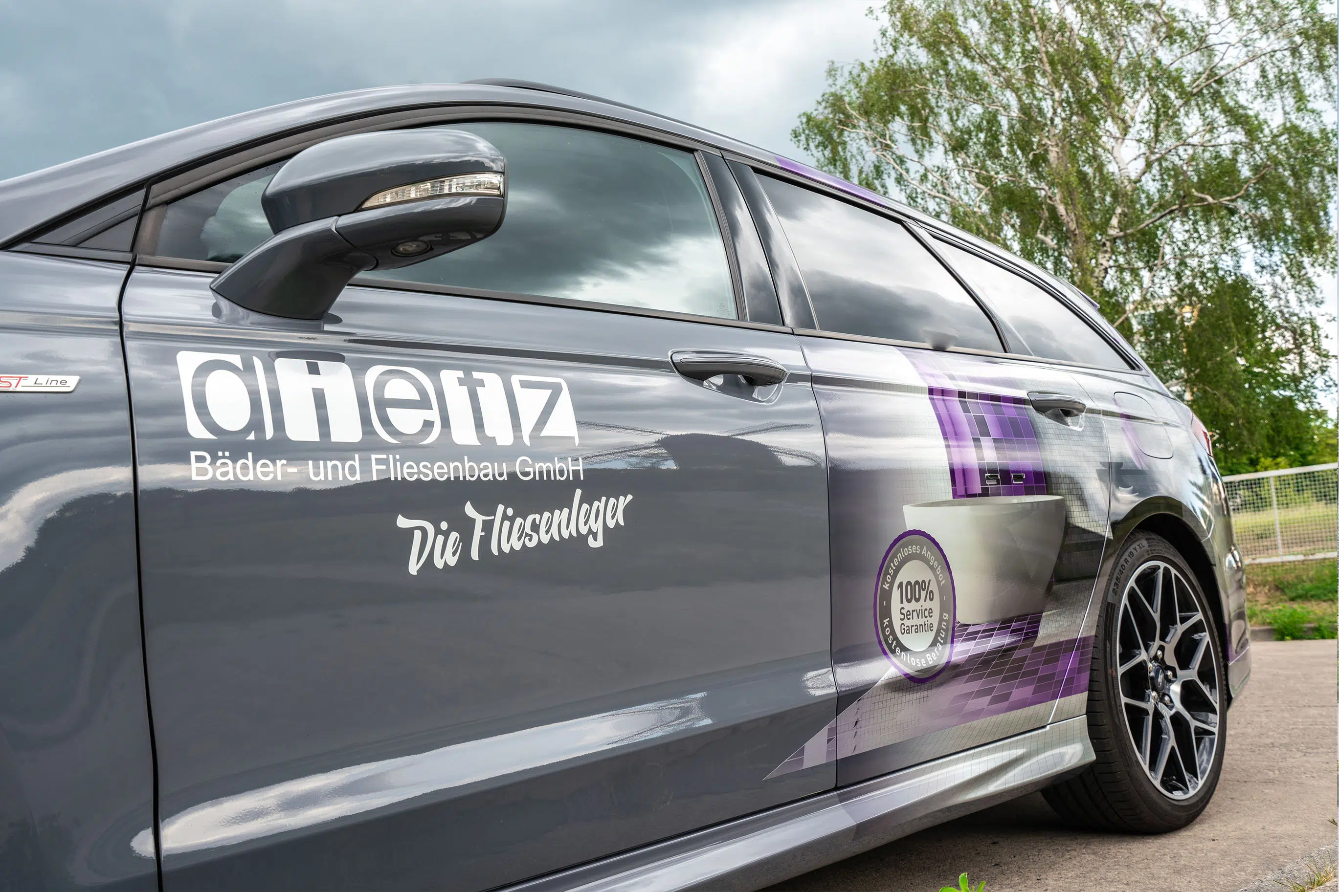 Dieses Bild zeigt ein Fahrzeug mit einer professionellen Werbefolierung für die Firma "Dietz Bäder- und Fliesenbau GmbH". Das Design kombiniert klare, weiße Schriftzüge mit einer grafischen Darstellung von Fliesen in Lila- und Grautönen. Besonders auffällig ist das runde Siegel mit der Aufschrift "100% Service Garantie". Die Gestaltung vermittelt einen hochwertigen und modernen Eindruck, passend zum Handwerk des Unternehmens.