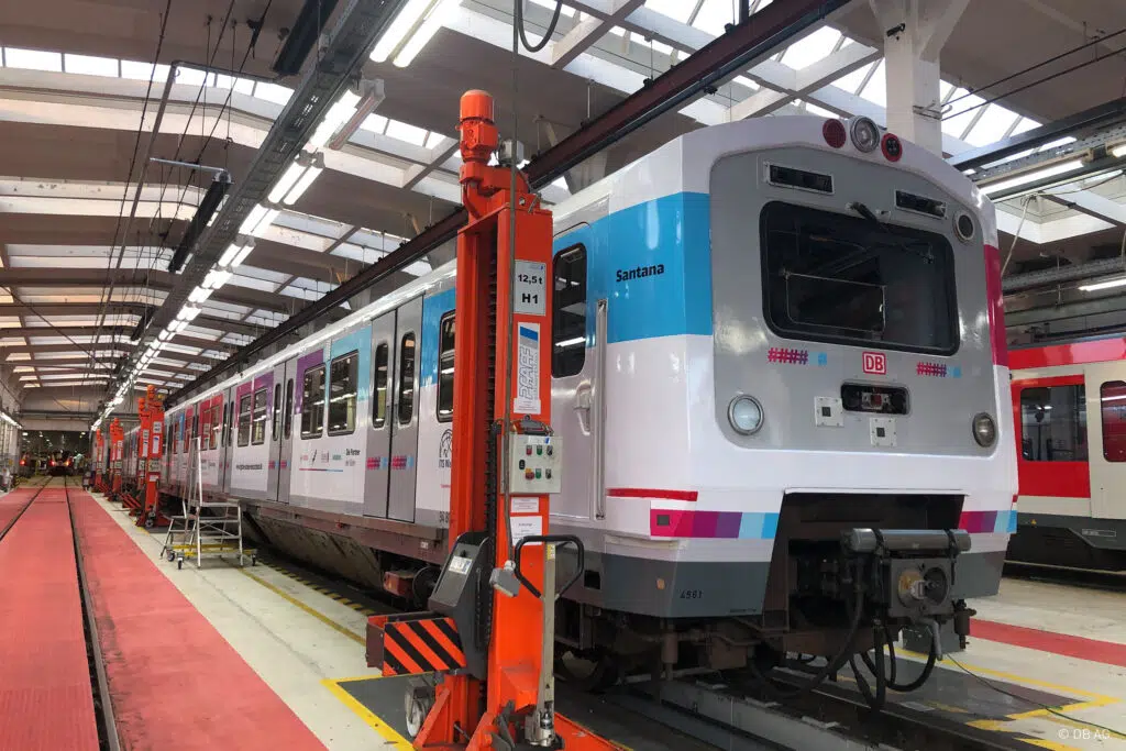 Das Bild zeigt einen modern gestalteten Zug in einer Werkstatthalle. Der Zug trägt eine helle Lackierung mit blauen und pinken Akzenten sowie das DB-Logo. Eine orange Hebevorrichtung steht im Vordergrund. Im Hintergrund sind weitere Züge und Werkstatteinrichtungen zu sehen.