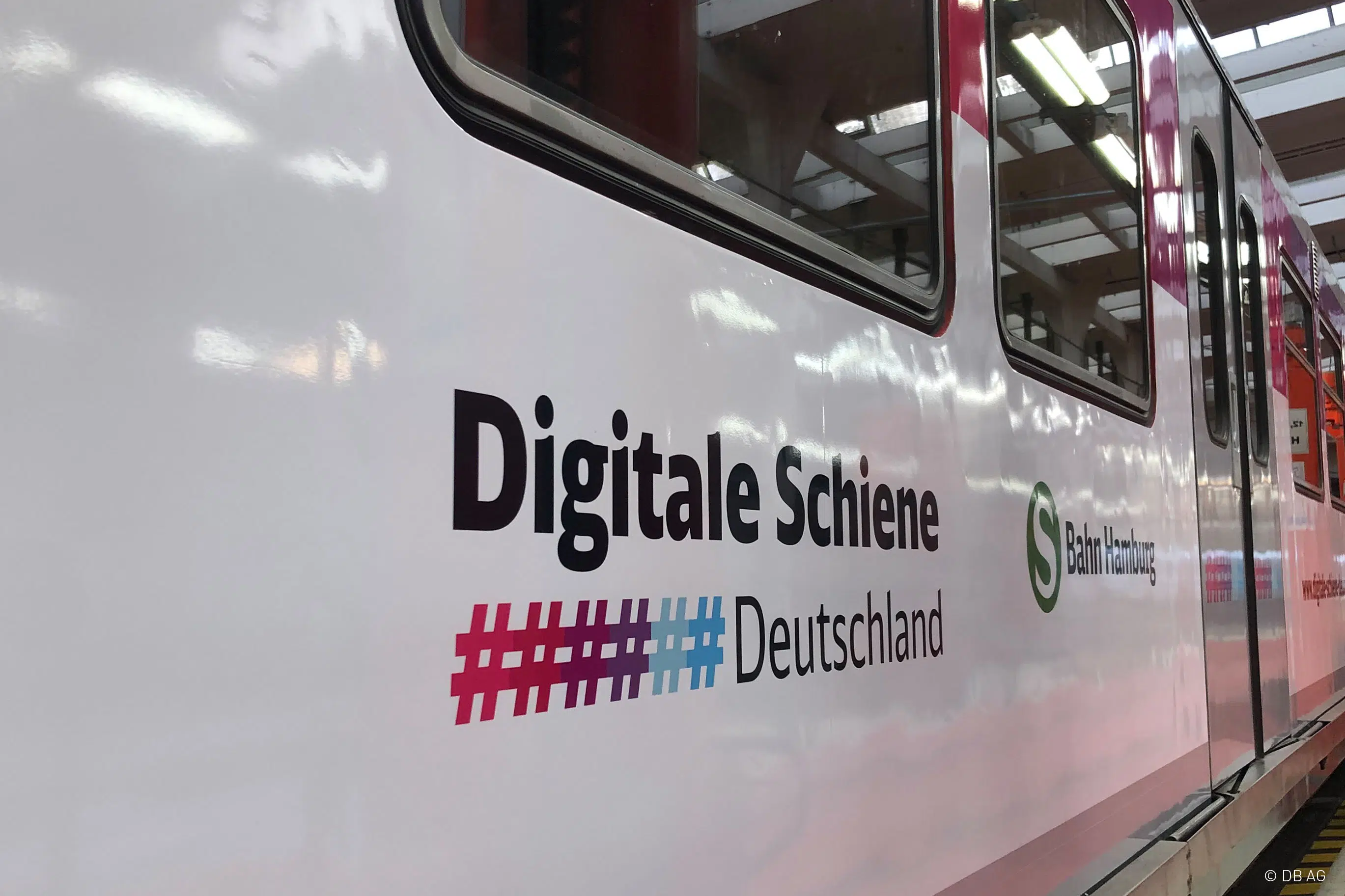 Das Bild zeigt die Außenansicht eines Zuges mit weißer Lackierung und der Aufschrift „Digitale Schiene Deutschland“. Darunter befindet sich ein mehrfarbiges Hashtag-Symbol. Zudem ist das Logo der S-Bahn Hamburg zu sehen. Die Fenster spiegeln die Hallenbeleuchtung.