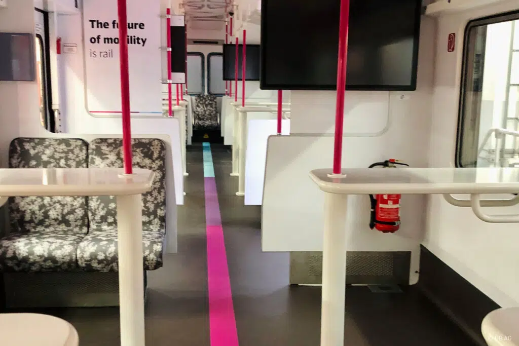 Das Bild zeigt den modernisierten Innenraum eines Zuges mit hellen Sitzen, weißen Tischen und pinken Haltestangen. Ein bunter Streifen verläuft mittig auf dem Boden. An den Wänden befinden sich Bildschirme und Beschriftungen, darunter „The future of mobility is rail“.