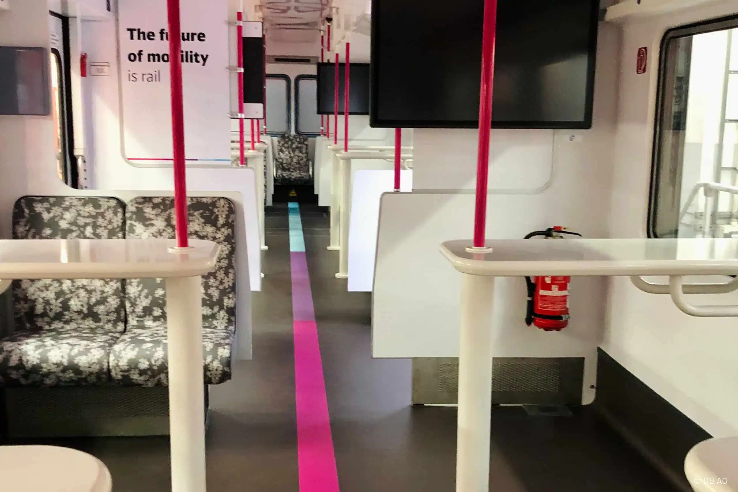 Das Bild zeigt den modernisierten Innenraum eines Zuges mit hellen Sitzen, weißen Tischen und pinken Haltestangen. Ein bunter Streifen verläuft mittig auf dem Boden. An den Wänden befinden sich Bildschirme und Beschriftungen, darunter „The future of mobility is rail“.