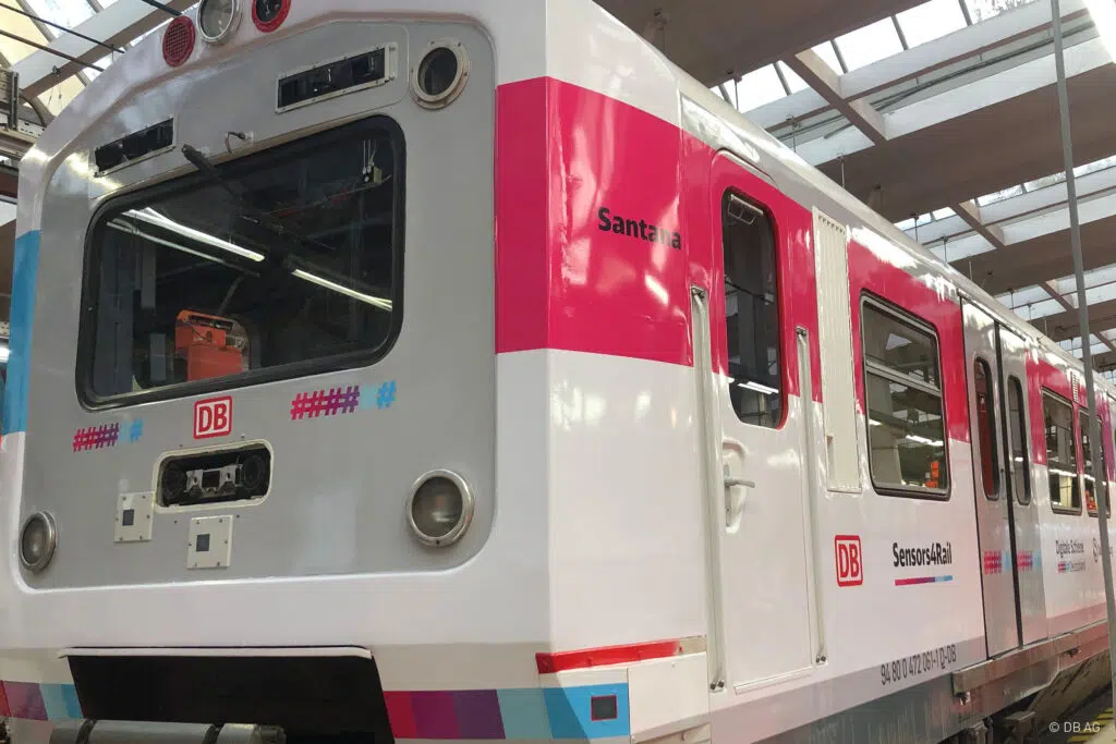 Das Bild zeigt einen modern folierten Zug der Deutschen Bahn im Innenraum eines Wartungsdepots. Die Folierung kombiniert weiße, graue und magentafarbene Elemente mit dem Schriftzug „Santana“ und „Sensors4Rail“. Der Zug trägt das DB-Logo und reflektiert das Licht der Hallendecke.