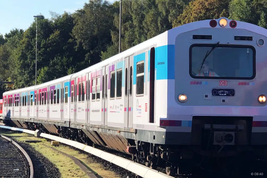 Das Bild zeigt einen modern folierten Zug der Deutschen Bahn auf einer kurvigen Gleisstrecke. Das Design enthält weiße, blaue und magentafarbene Elemente. Ein Lokführer mit Warnweste ist im Führerstand sichtbar. Im Hintergrund befinden sich Bäume und ein Bahnsignal.